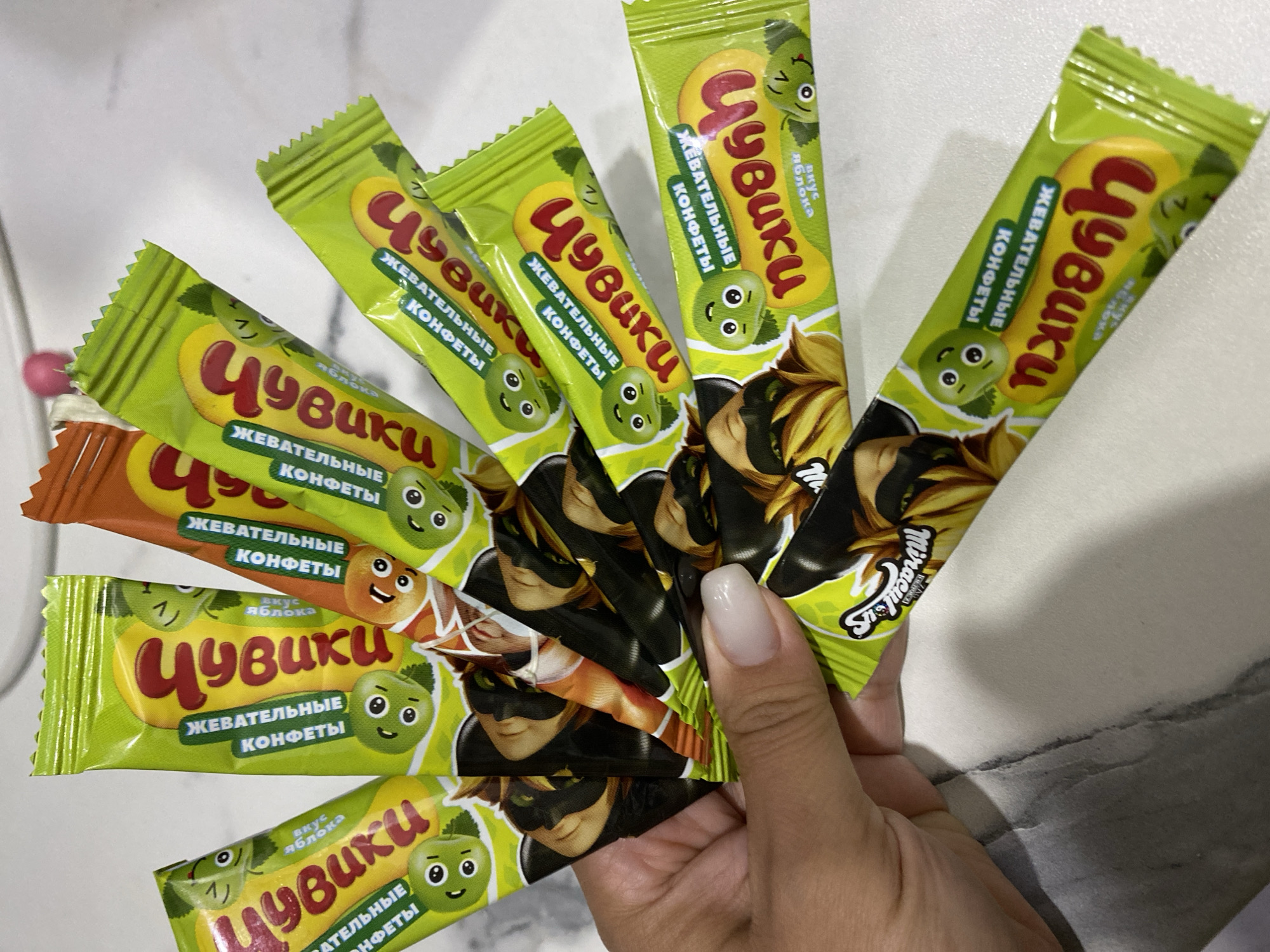 A hand with a pale pink nail is holding a cluster of individually wrapped candies. The candies are mostly green and yellow with red lettering. Some of the wrappers also feature cartoon characters that appear to be green, round fruits with eyes. One wrapper has a character that resembles a black cat with yellow hair. The background is a white, textured surface, possibly a countertop or table, with some grey marbling. The scene is an indoor setting, and there is no discernible information to suggest a specific location in the city of Ufa, Russia.