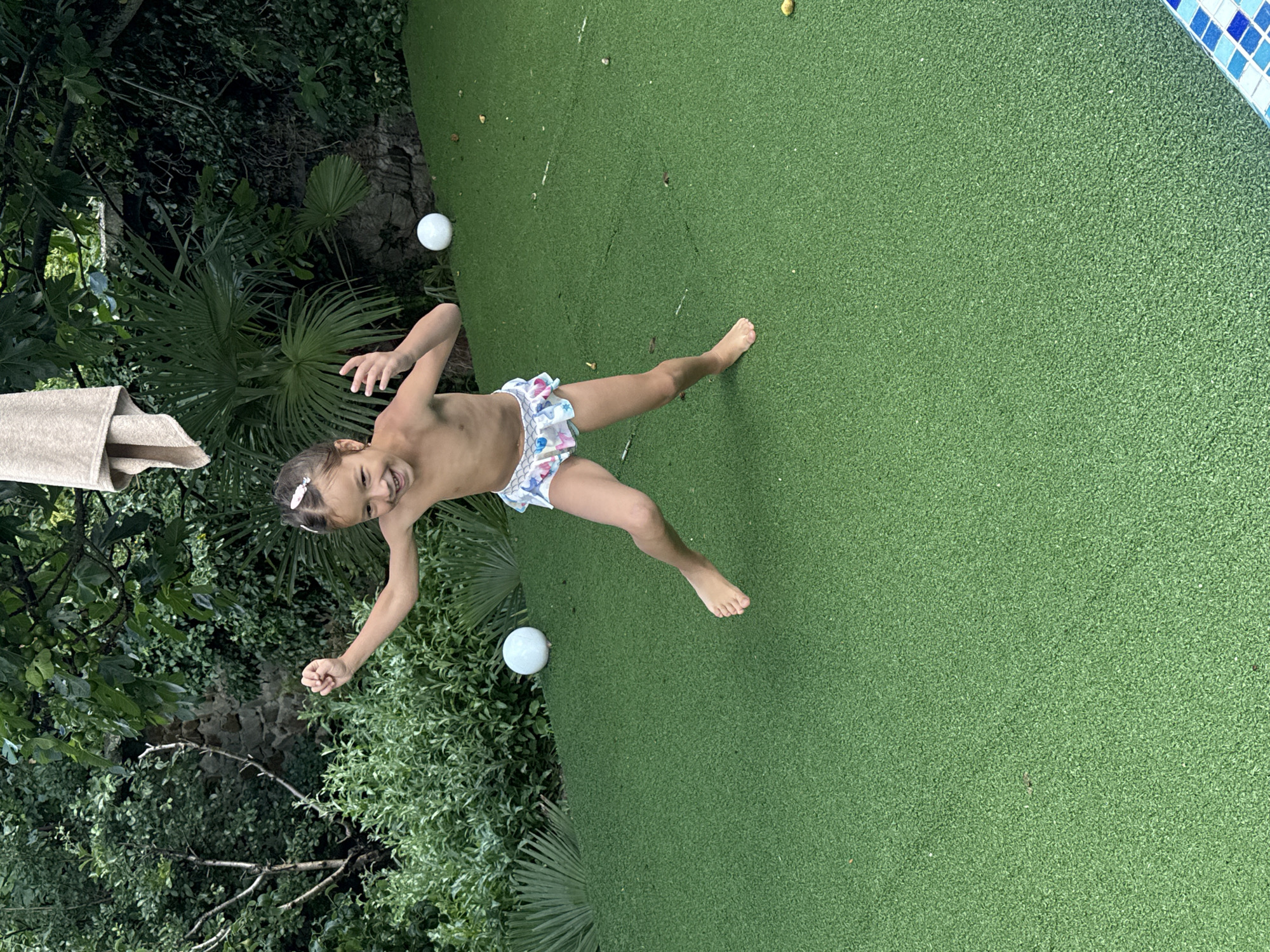 A young child, likely a girl, is captured mid-jump on a bright green, artificial turf surface. She appears to be topless, wearing only patterned swim shorts. Her arms are outstretched, and her body is contorted as if in the middle of a playful leap. A white hair accessory is visible in her dark hair, and her face is turned towards the camera with a wide, joyful smile and her eyes are closed.
The setting is outdoors, with lush green foliage and palm-like plants framing the left side of the image. To the right, a sliver of a pool border with blue and white square tiles is visible, suggesting the turf might be a poolside area. Two white, spherical objects, possibly balls, are scattered on the turf in the background, one near the foliage and another further away. The lighting suggests it's daytime, with the bright green of the turf indicating good weather. The overall mood is one of fun and carefree childhood activity. There is no visible text in the image.