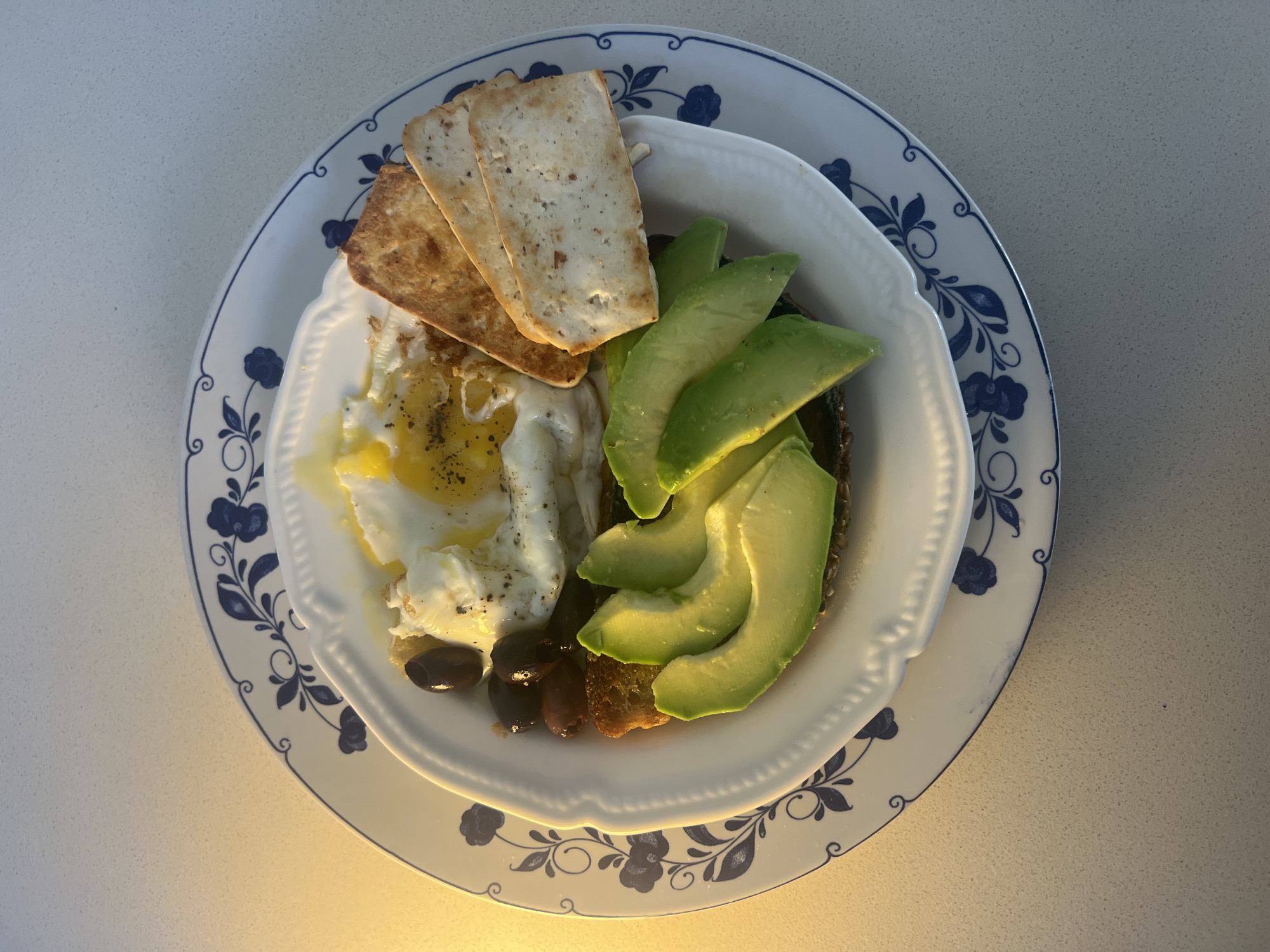 An overhead shot presents a well-arranged single serving of breakfast or brunch, prepared in Kiryat Bialik, Israel. The meal is served in a white, round bowl with a decorative scalloped edge, which rests on a larger white plate featuring a blue floral and scroll pattern along its rim.
The dish comprises several components: a perfectly cooked sunny-side-up fried egg, liberally seasoned with black pepper, is positioned on the left side of the bowl. Next to the egg are a few dark, whole olives, likely pitted. On the opposite side, several vibrant green slices of fresh avocado are neatly fanned out, seemingly placed on a piece of dark, possibly whole-grain toast or bread which is largely obscured. Three rectangular, light brown crackers or crispbread pieces are stacked near the top of the egg.
The entire arrangement is set on a light-colored, subtly textured countertop. The lighting appears to be ambient indoor light, suggesting a daytime setting. There is no visible text in the image.