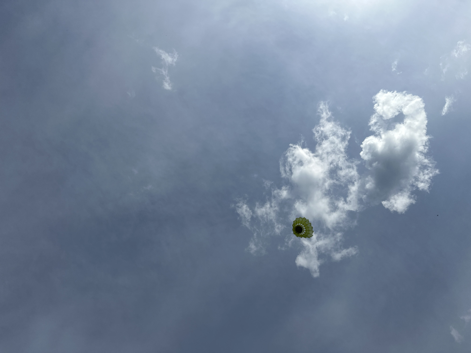 The image shows a low-angle view of the sky. The sky is overcast with a mix of grey and white clouds. A green and yellow shuttlecock is suspended in the air, seemingly hanging in front of the clouds. No people or discernible buildings are visible. There is no information suggesting the city of Nizhny Novgorod, Russia.