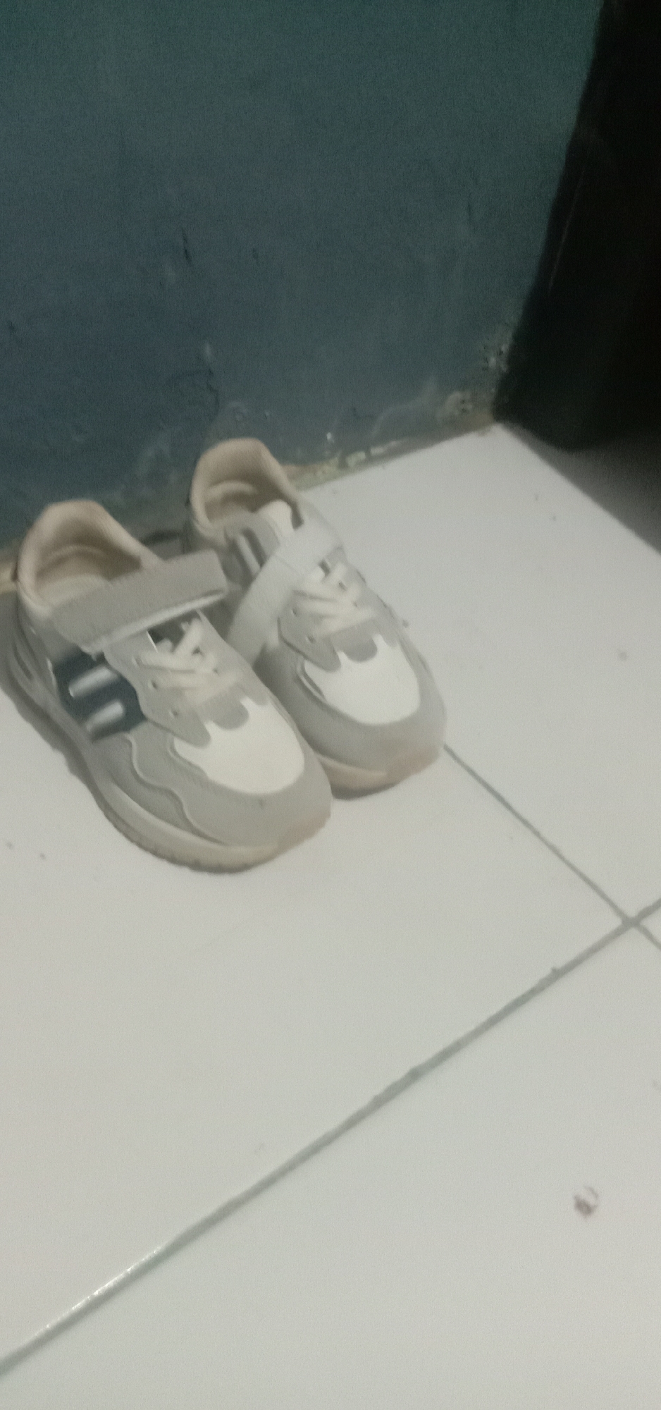 A pair of light-colored athletic shoes, likely for children due to their small size and velcro straps, are placed on a tiled floor. The shoes have a predominantly white and light gray color scheme with some darker gray or black accents on the sides of the left shoe. They are positioned near a wall that appears to be painted a dark, muted color, possibly dark blue or teal. The lighting in the scene is dim, casting some shadows and creating a slightly hazy or blurry effect. The tiled floor is light-colored, with visible grout lines. This scene is likely located in a residential setting within the city of Kendari, Indonesia, perhaps in a hallway or entryway.