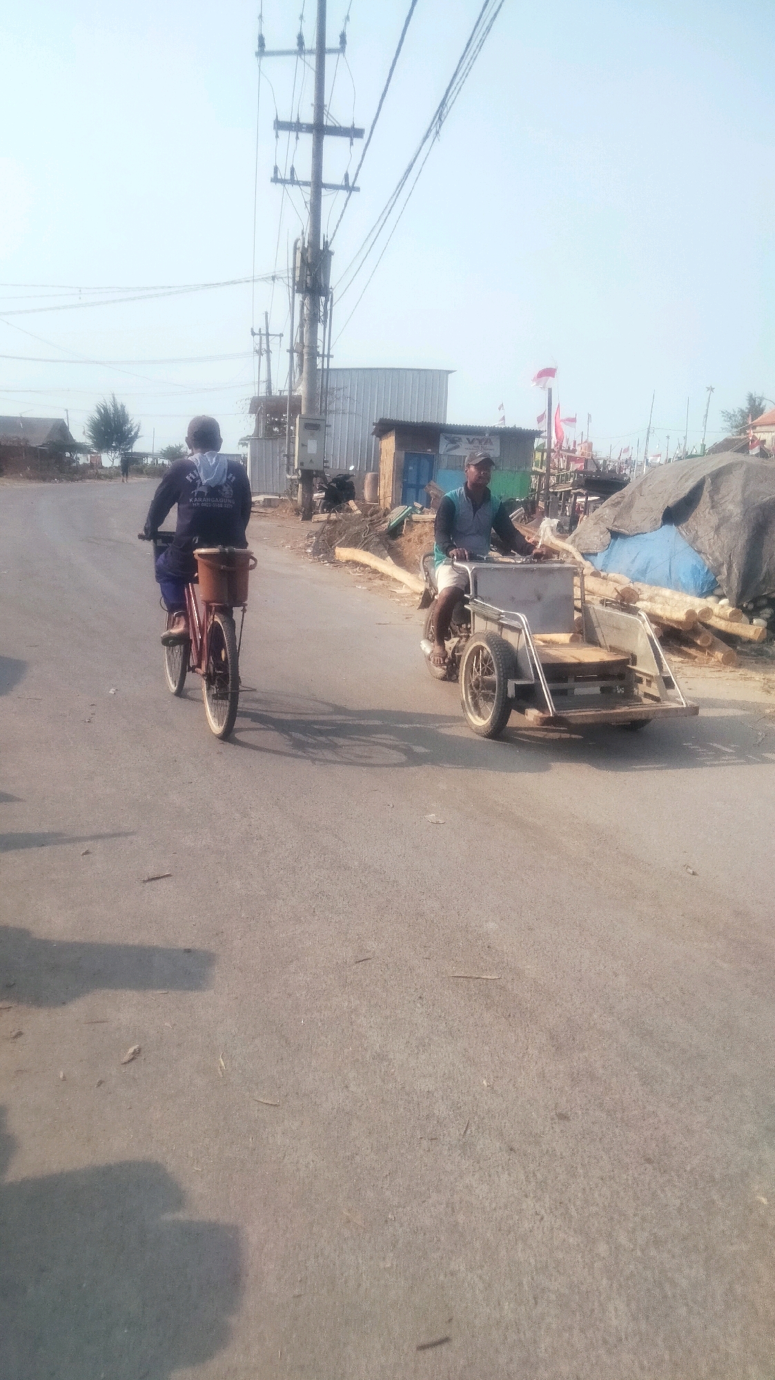 In Palang, Indonesia, on a bright day, two individuals are seen on a paved road with a backdrop of utility poles and buildings. To the left, a person is riding a red bicycle, facing away from the camera, with a basket attached to the rear. To the right, a man is operating a motorized tricycle with a sidecar, also moving forward on the road. In the background, there are several structures, including a tin-roofed building, a smaller wooden shed, and a collection of large wooden logs, some covered with tarps. Red and white flags are visible on poles in the distance, suggesting a possible local event or celebration. The overall scene depicts a typical day on a road in a coastal or semi-rural Indonesian town.