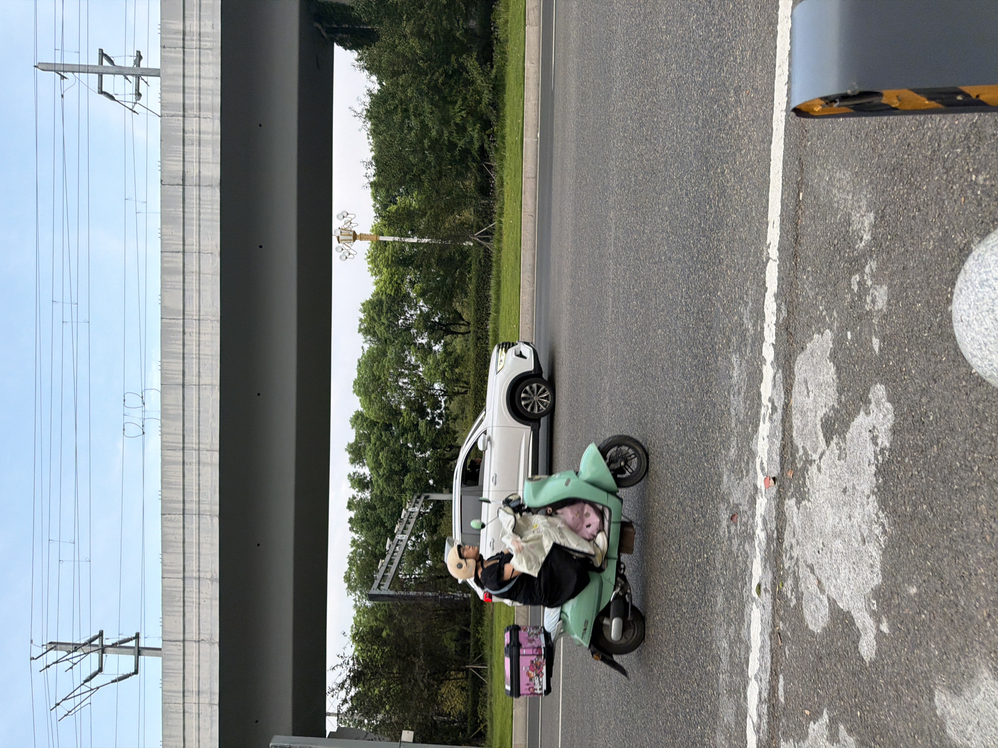 In Jiangjin, China, a person wearing a helmet and a black dress rides a mint green scooter on a paved road. The scooter has a pink and white patterned storage box attached to the rear. A white SUV is visible behind the scooter. Overhead, a concrete overpass runs across the image, with power lines and utility poles visible against a cloudy sky. Trees and greenery line the side of the road. The foreground of the image shows the asphalt road surface with white markings and some debris.