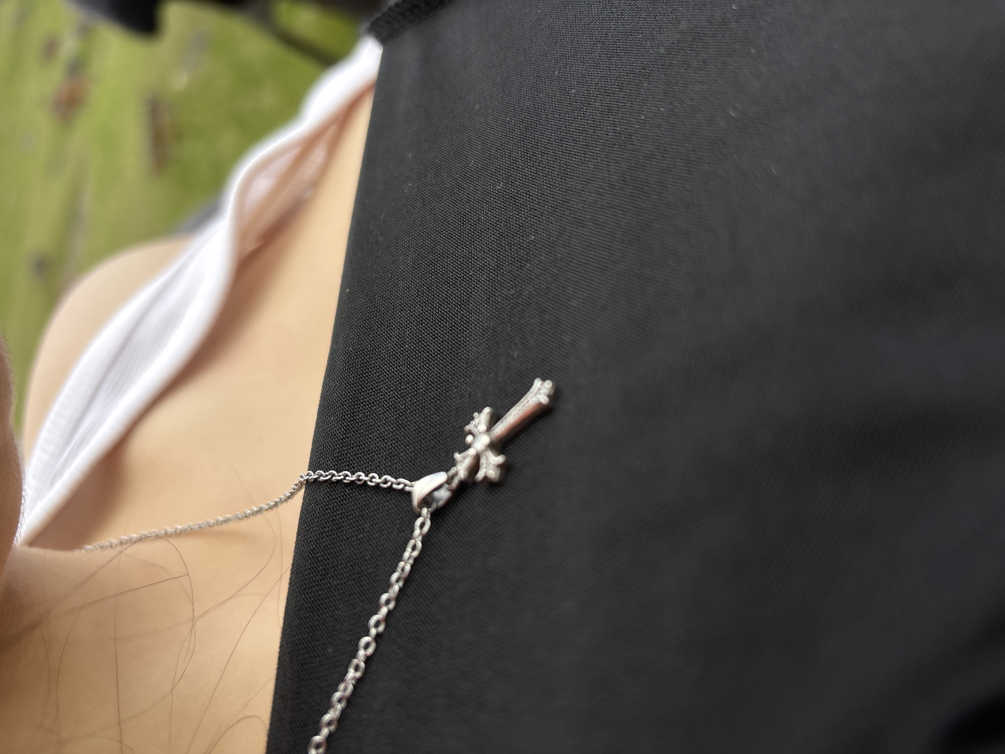 A close-up shot depicts a person wearing a black garment with a silver cross pendant necklace. A white undergarment is also visible. The background is blurred and out of focus, showing green hues suggesting foliage or grass. There is no indication of the location being Tlik, Armenia.