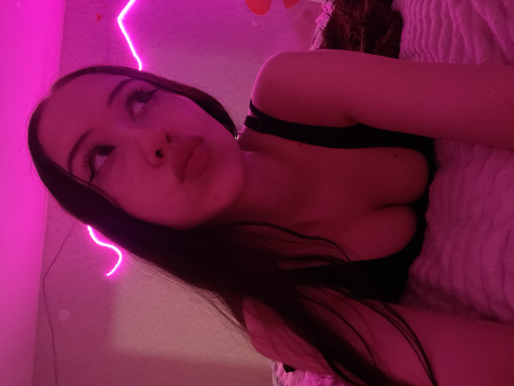 The image shows a young woman with long, dark hair, wearing a black top. She is lying on a textured surface, likely a bed, and is illuminated by pink neon lighting. The background is a pink wall with a neon light strip creating a curved shape. The woman is looking away from the camera with her lips pursed. The scene appears to be indoors, possibly a bedroom. There is no information in the image that can determine the location in Votkinsk, Russia.