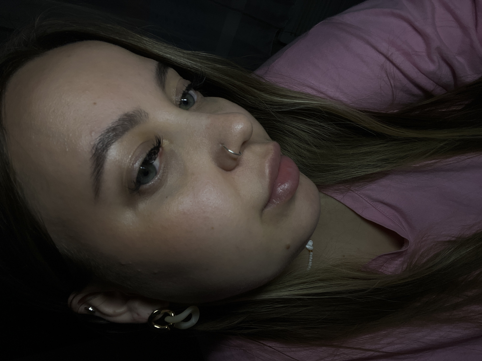 A close-up, dimly lit photograph shows a person with blonde hair lying down, presumably indoors, in what appears to be Novopokrovskaya, Russia. The person is wearing a pink shirt and has a nose ring, earrings, and a beaded necklace. Their eye color appears to be grey. Details about the location or specific actions are difficult to determine due to the image's composition and lack of environmental context.