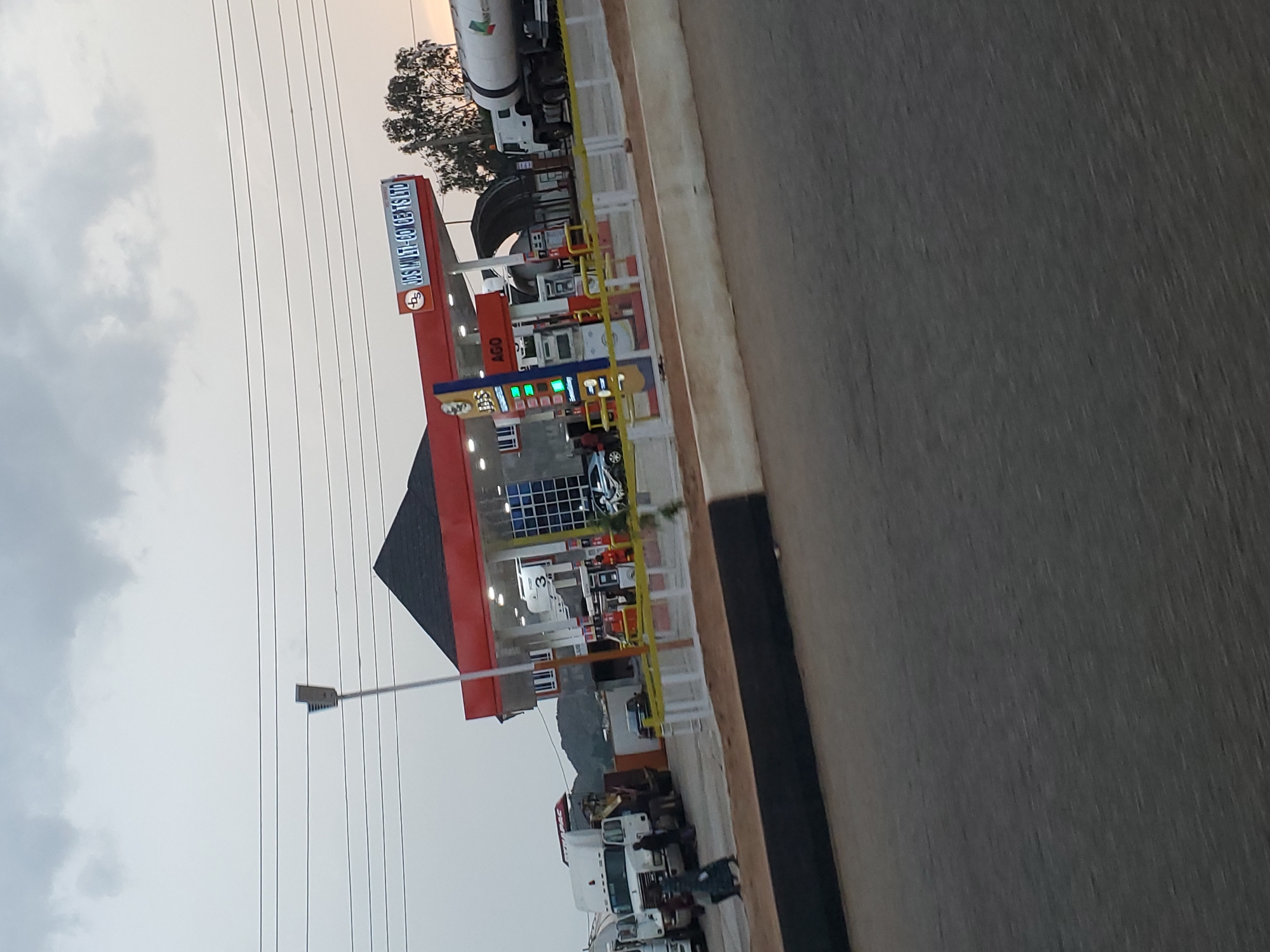The image shows a view of a gas station in Bukuru, Nigeria. The gas station is branded "JDS MULTI-CONCEPTS LTD" and has "AGO" displayed on one of its pumps. Several trucks are visible at the gas station, including a white tanker truck. Some people are also present, including a person standing near one of the trucks and others near the gas pumps. The sky is cloudy, and it appears to be either early morning or late evening. The gas station has a modern design with a red and white canopy over the pumps. A street lamp is visible in the upper left quadrant of the image.