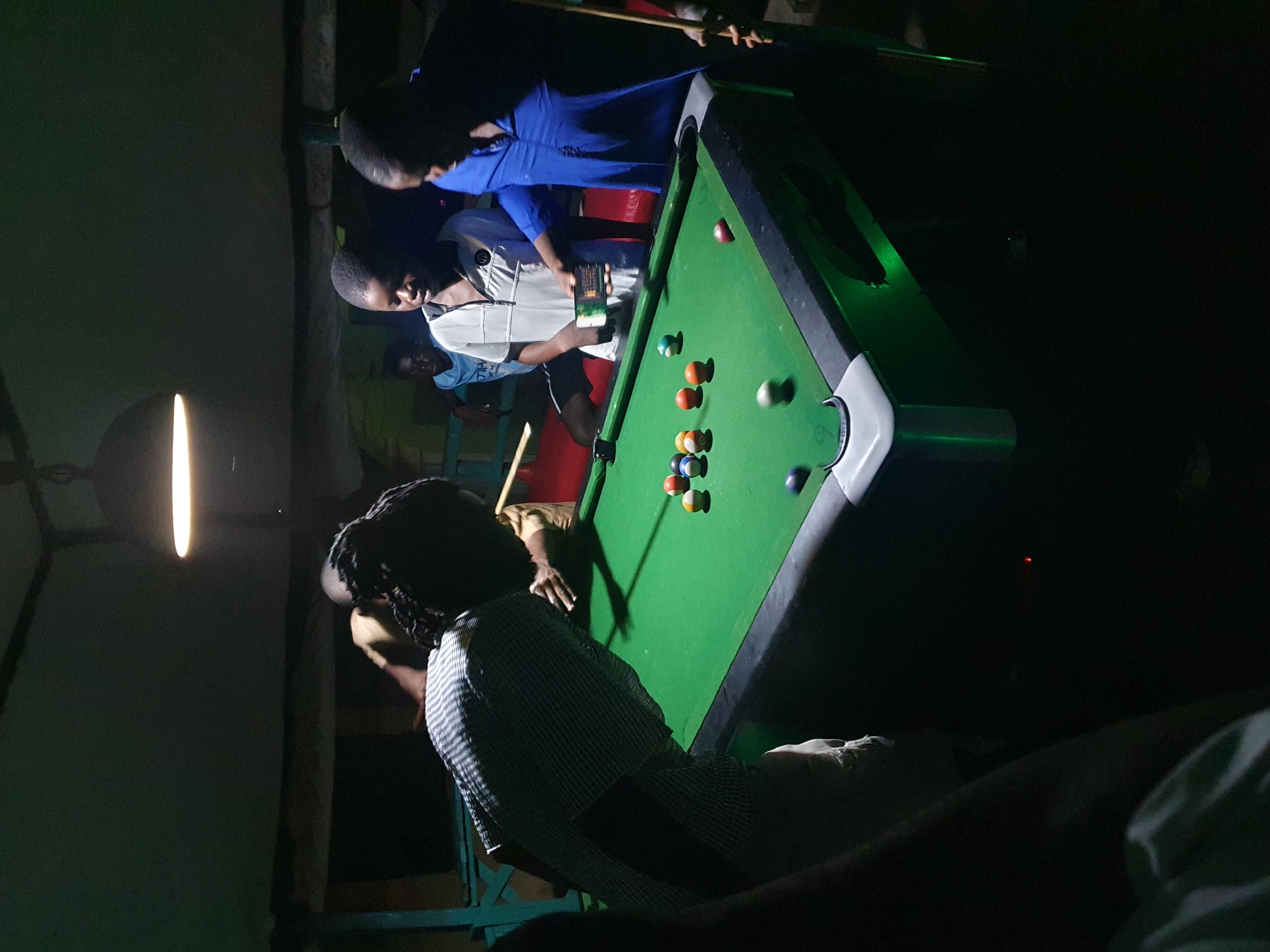 This is a nighttime, indoor photograph of a pool table game in progress. Five individuals are visible. A person wearing a black and white striped shirt is leaning over the pool table, preparing to take a shot. To their right, a boy in a white shirt is looking at a smartphone, and to his right, another person in a blue shirt is observing. In the background, two other boys are standing near the pool table. The pool table itself is green with several colorful balls on it. The environment appears to be a dimly lit recreational space with a visible ceiling and a hanging lamp. The image seems to have been taken in Lokoja, Nigeria, given the context provided.