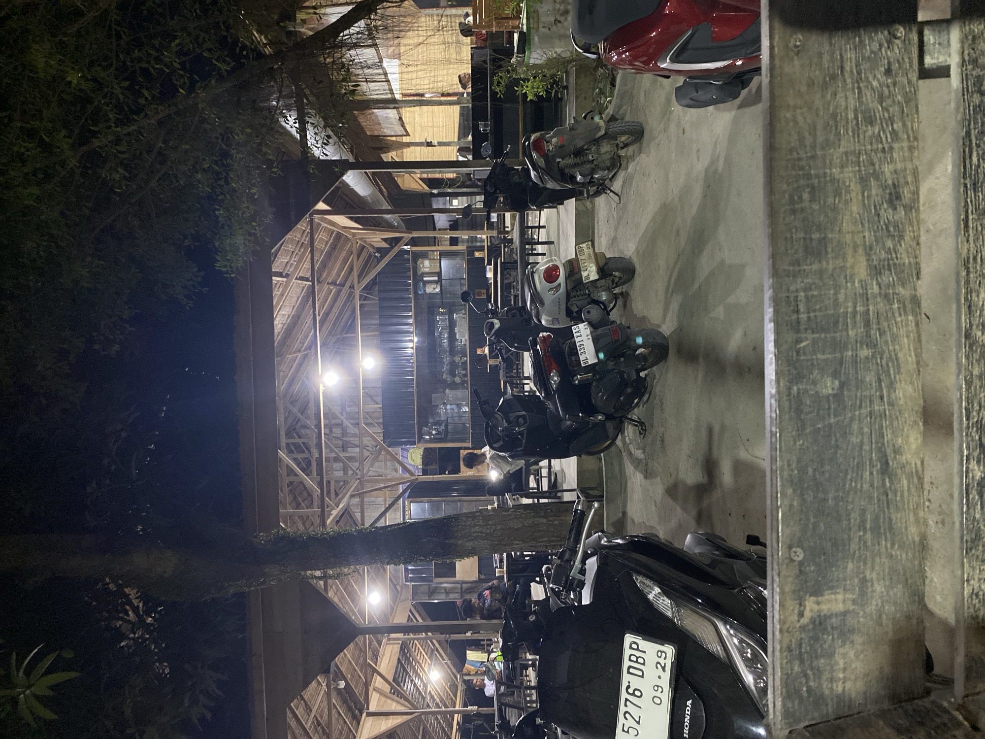 The image captures an evening scene at an outdoor or semi-outdoor establishment, likely a cafe or restaurant, in Seuneubok Teungoh, Indonesia. The foreground and mid-ground are occupied by numerous parked motorcycles and scooters on a concrete surface. In the immediate foreground, a sturdy wooden barrier partially obscures a black Honda scooter with license plate '5276 DBP', showing an expiry date of '09-29'. Other visible scooters include a white one with license plate 'BL 8391 ZAS' (expiry '12-25') and another white one displaying 'BL 4117 OBP' (expiry '06/25'). A red scooter is also visible on the far left.
The main structure, brightly lit by artificial hanging lamps, features a distinctive wooden frame and what appears to be a thatched or bamboo roof, providing a rustic aesthetic. Inside, several individuals are visible, seated or standing, suggesting patrons socializing or dining. A tall tree trunk ascends through the middle of the frame, with dense green foliage from surrounding trees prominent on the left side and upper portions of the image, emphasizing the outdoor setting. The presence of 'BL' on the license plates further situates the scene in Aceh province, Indonesia. The overall atmosphere suggests a lively night spot.