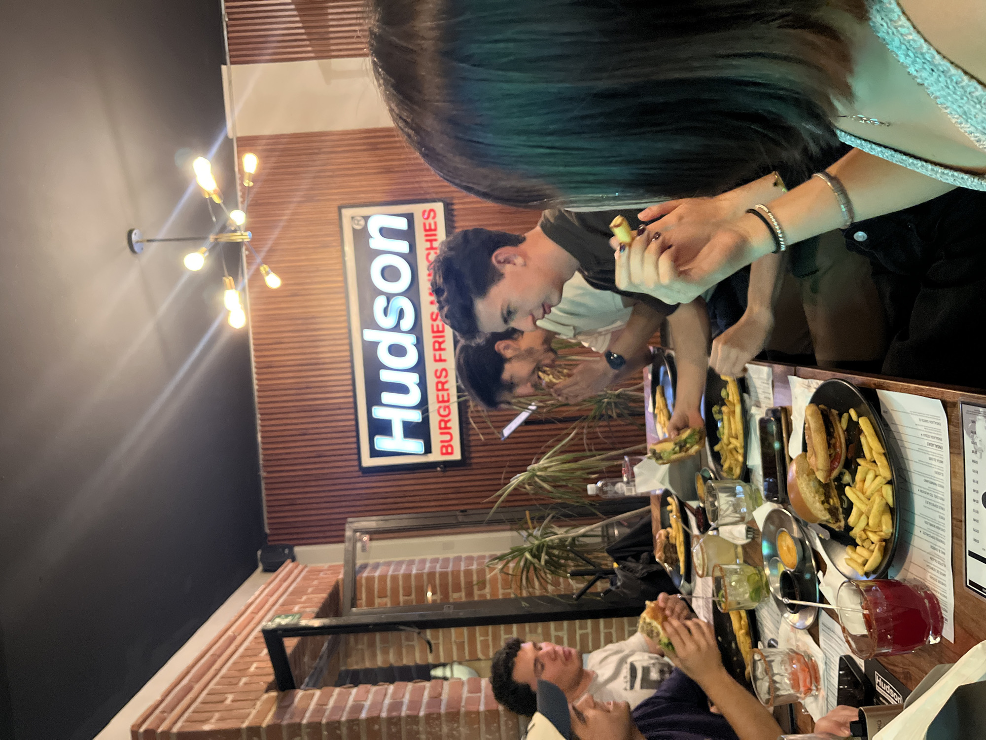 The media file captures an indoor scene at a burger restaurant, likely "Hudson," in San Andrés Cholula, Mexico, during the evening. Several young adults are gathered around wooden tables, eating and socializing.
In the foreground, the back of a woman's head with dark hair is visible. Her arm is extended towards the table, holding a French fry between her fingers, and she wears multiple bracelets on her wrist. Across from her, a young man with dark hair is seen looking down at his food. Further down the table, other individuals are also visible, engaged in eating.
The tables are laden with plates of food, prominently featuring burgers and generous servings of golden-brown French fries. Various drinks are also present, including clear glasses with lime slices and a red beverage in a handled mug. The restaurant's interior design includes walls with vertical wooden paneling and sections of exposed reddish-brown brick. A modern light fixture with multiple illuminated bulbs extends from a dark wall, providing ambient lighting.
A rectangular sign with a white background and black border is mounted on the wooden-paneled wall, displaying the word "Hudson" in large, prominent lettering. Below it, smaller white text reads "BURGERS FRIES & MUCHIES" (likely "MUNCHIES"). The scene conveys a relaxed and casual atmosphere, typical of a burger joint.