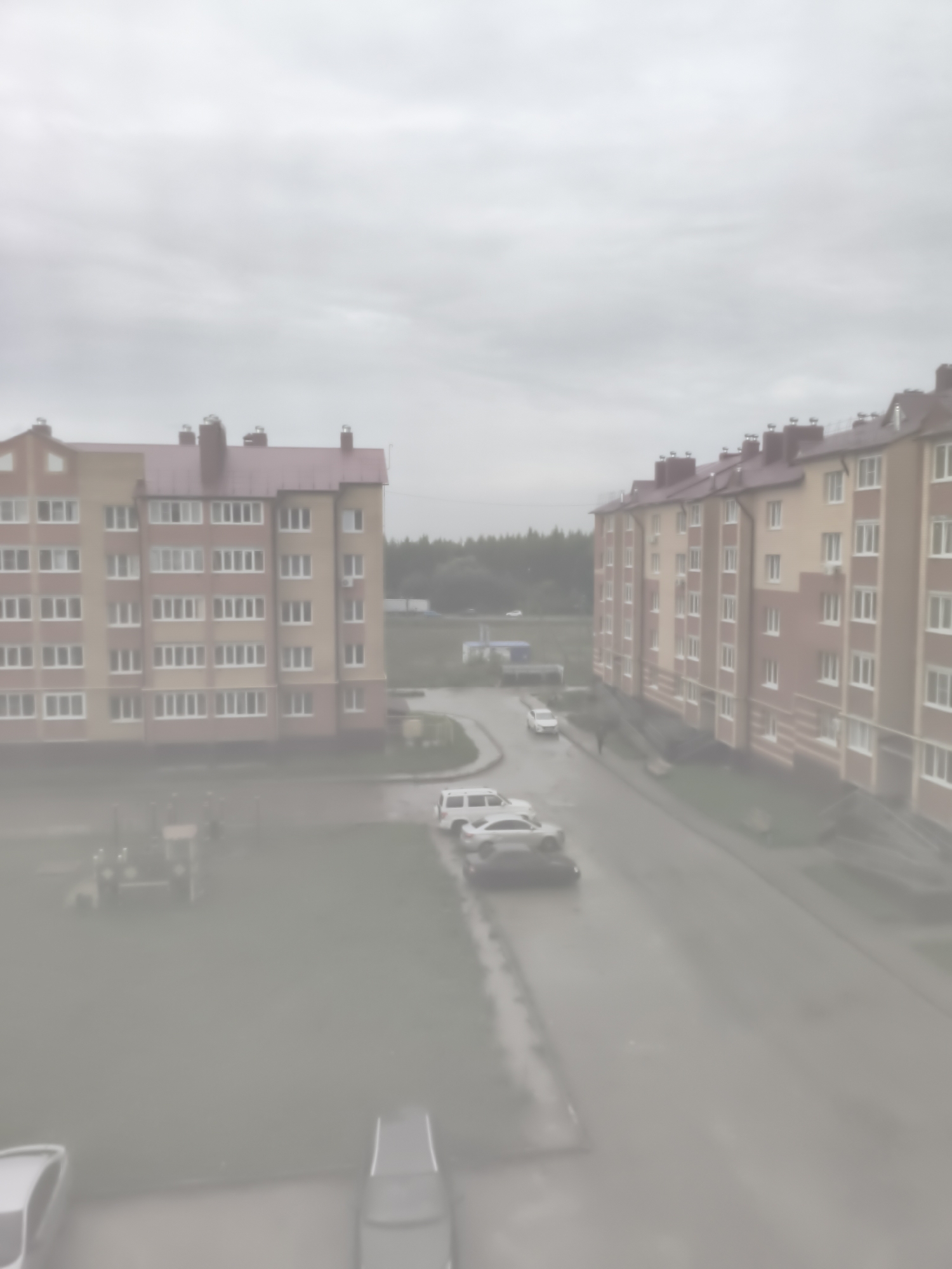 The image depicts an overcast day in the city of Tsivilsk, Russia, with a view of residential apartment buildings and a street. On the left, a light yellow and brick-red apartment building stands with a red roof. To its right, a similar apartment building, also with a red roof, stretches along the street. Several cars are parked on the street and on a patch of grass in front of the buildings. A playground-like structure with a tractor toy is visible on the grass. The street appears wet, suggesting recent rain. The overall atmosphere is subdued due to the cloudy weather.