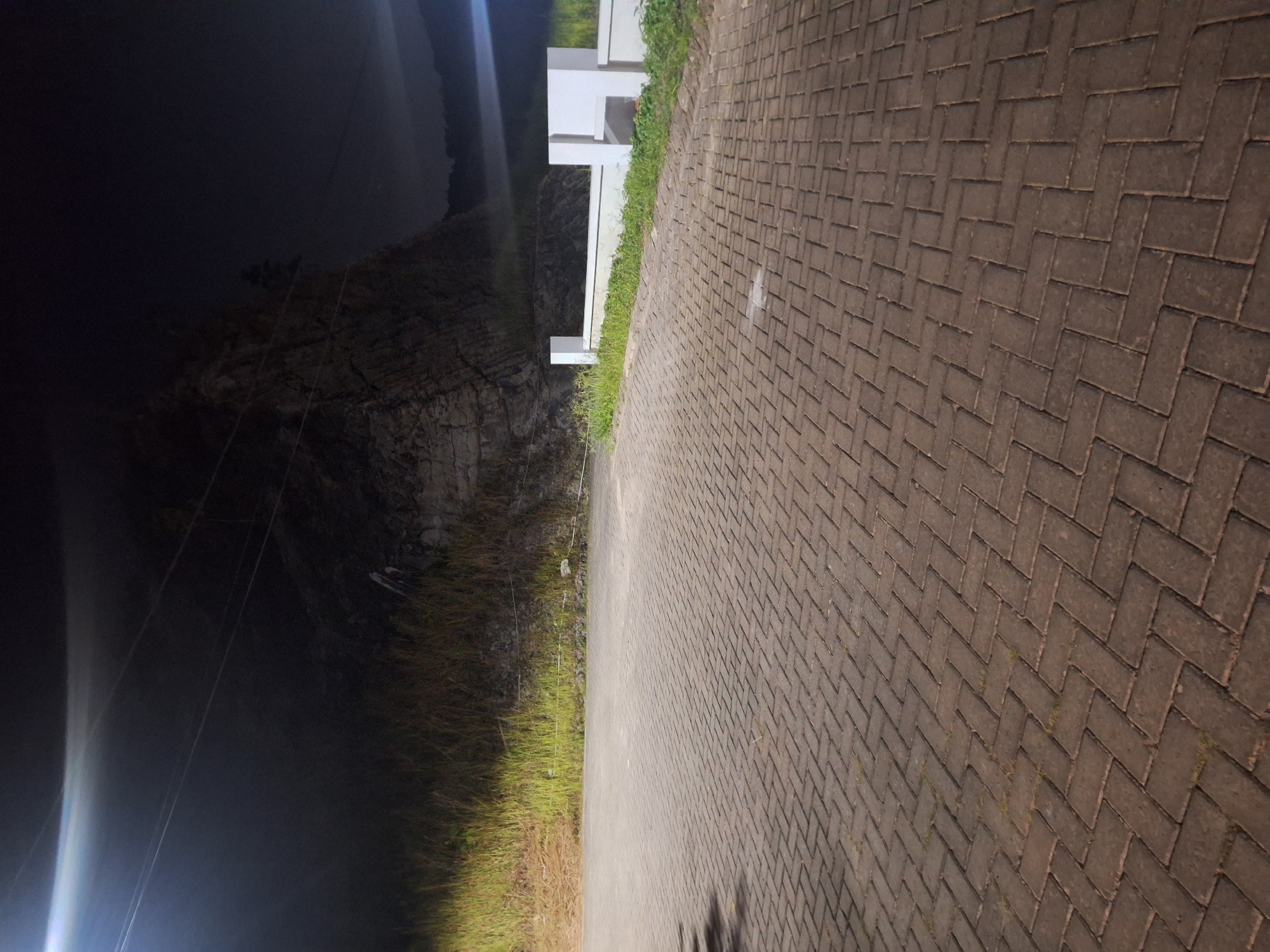 The image depicts a paved area at night in Samarinda, Indonesia. The foreground is dominated by a herringbone pattern of reddish-brown paving stones, with patches of grass and weeds growing between them. To the right, a white concrete wall with vertical posts is visible, bordering a grassy area and the base of a rocky cliff face. The cliff is dark and textured, with some vegetation clinging to it. Overhead, power lines are strung across the sky, illuminated by a bright light source casting a beam downwards and to the left, creating a hazy effect in the dark. The overall atmosphere is dimly lit, with artificial lighting providing the primary illumination.