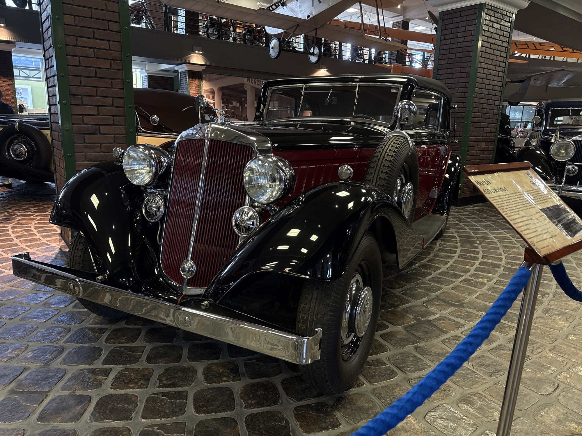 In the city of Arkhangel’skoye, Russia, at the Arkhangelskoe Estate Museum, a black and maroon vintage convertible automobile is displayed indoors on a cobblestone floor. The car, identified by a plaque as a "Horch 830 cabriolet", has a chrome grille, large round headlights, and a spare tire mounted on the driver's side. The interior of the museum is visible, with brick pillars, exposed ceiling beams, and what appear to be airplane exhibits suspended overhead. A blue velvet rope barrier separates the viewer from the vehicle. No people are prominently featured.
