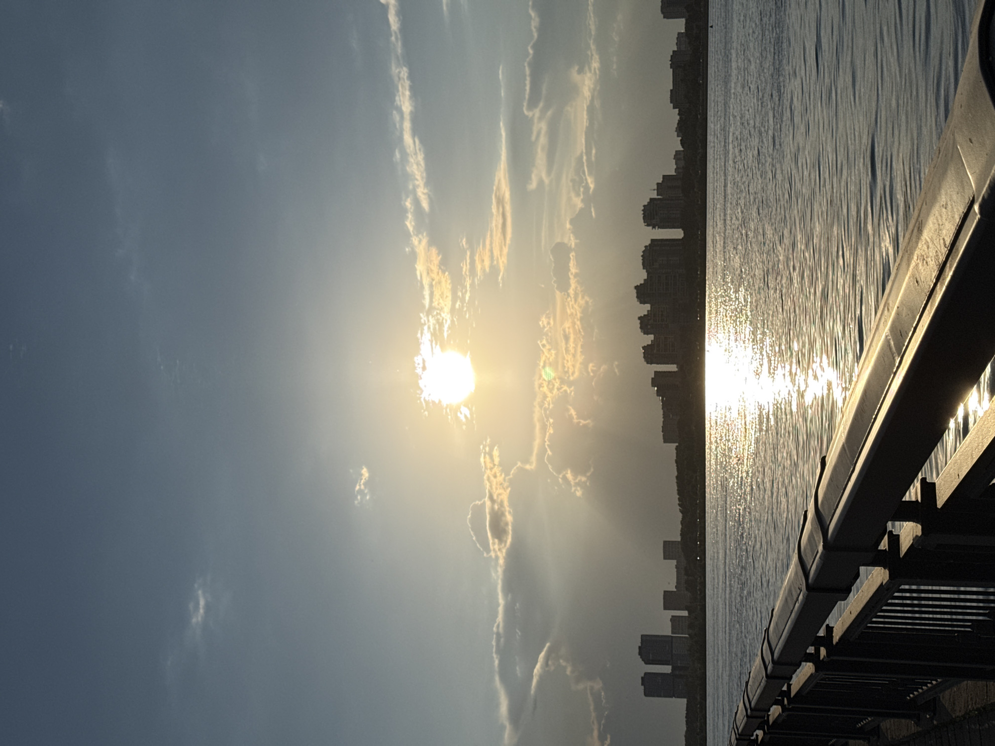 The image shows a waterfront scene in Kazan, Russia. The sun is positioned in the sky, partially obscured by clouds, with light rays emanating outwards. Reflected sunlight glitters on the surface of the water. Along the water's edge, the silhouettes of buildings, characteristic of the city skyline, can be discerned. A portion of a metal railing or structure is visible in the foreground.