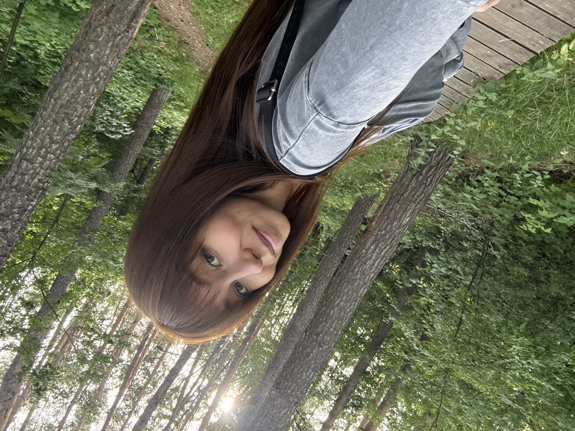 The media file is a portrait-oriented, low-angle selfie of a young woman with long, dark brown hair and bangs. She is looking towards the camera with a neutral expression. Her eyes are green, and she has fair skin. She is wearing a grey hooded jacket with a black strap across the chest, possibly from a bag.
The woman is outdoors, in what appears to be a forest or wooded area. Several trees are visible, with rough bark and green leaves. Sunlight filters through the trees, suggesting it is daytime, likely late afternoon or early morning due to the angle of the light. There is also a wooden boardwalk or path visible in the background, with green grass and other vegetation.
The overall impression is one of a casual, spontaneous moment captured in a natural setting. The orientation of the image is unusual, as it appears to be taken with the camera held upside down. The location context provided is Chaikovsky, Russia. There is no visible text in the image.
