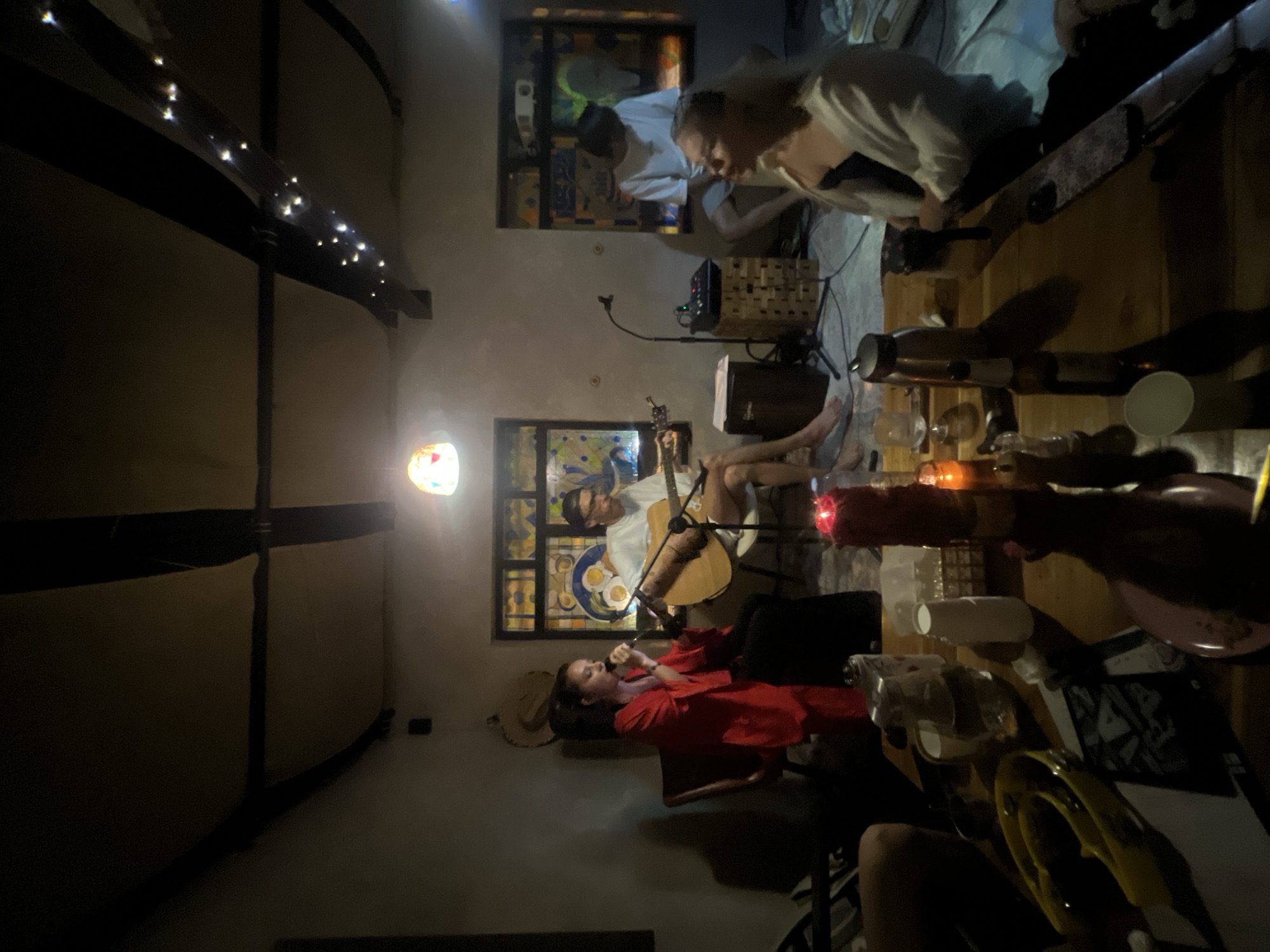 In Ko Samui District, Thailand, a dimly lit, rustic room hosts an intimate musical performance. A woman in a red robe stands near a microphone, singing, while a man in a white t-shirt plays an acoustic guitar. Another man in a white t-shirt sits on a stool, and a woman with blonde hair in a cream cardigan faces away from the camera. The room features stained-glass windows, exposed wooden beams decorated with fairy lights, and a chandelier. A wooden table in the foreground is laden with drinks, bottles, and a red candle, suggesting a casual gathering. The atmosphere is relaxed and convivial, capturing a moment of live music and social interaction.