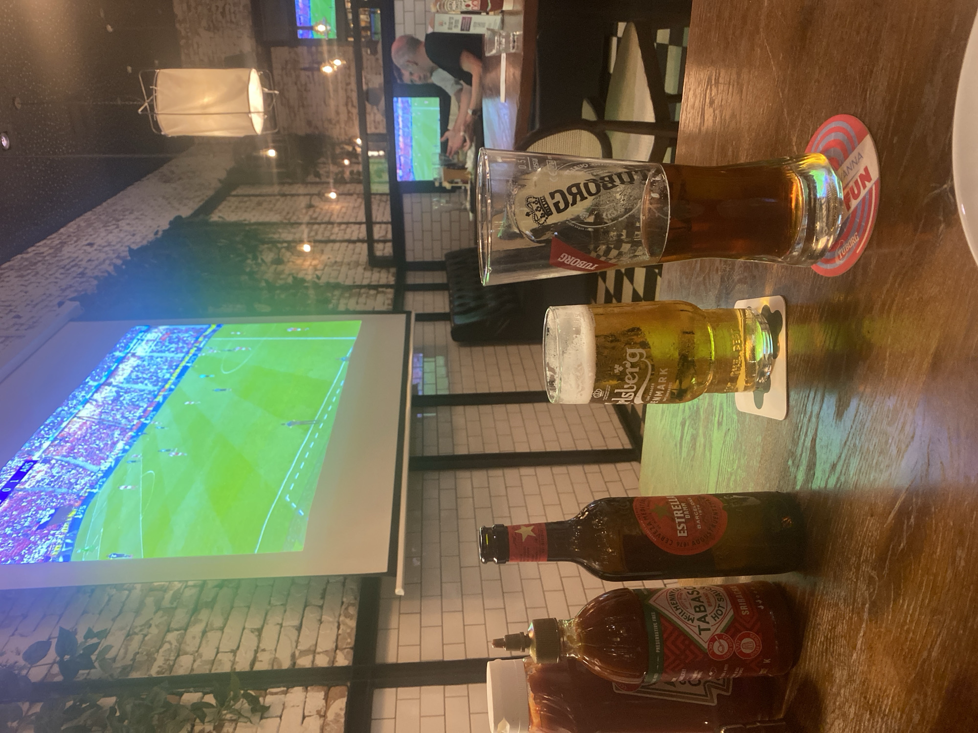 The scene is an indoor setting that appears to be a bar or restaurant, likely in Kiryat Bialik, Israel. A projector screen displays a soccer game, suggesting a sports-watching venue. Several alcoholic beverages are visible on a wooden table, including a glass of Tuborg beer, a glass of Carlsberg beer, and a bottle of Estrella Damm beer. Also present are bottles of Tabasco sauce and what appears to be sriracha sauce. In the background, another person is seen standing at a counter. The interior design includes exposed brick walls, a drop light fixture, and modern furniture. The overall atmosphere suggests a casual and social setting.