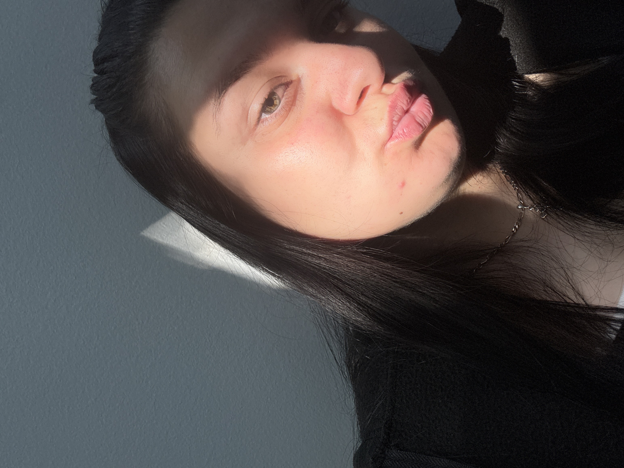 The image shows a close-up, low-angle selfie of a young woman with dark hair and fair skin. She is wearing a black top and a silver necklace. Her lips are pursed, and her head is tilted slightly. Sunlight casts a shadow across her face, highlighting her right eye and cheekbone. A light-colored wall is visible in the background, with a sharp diagonal shadow from an unseen object. The image appears to have been taken indoors. There is no indication of the location being the city of Plovdiv, Bulgaria.