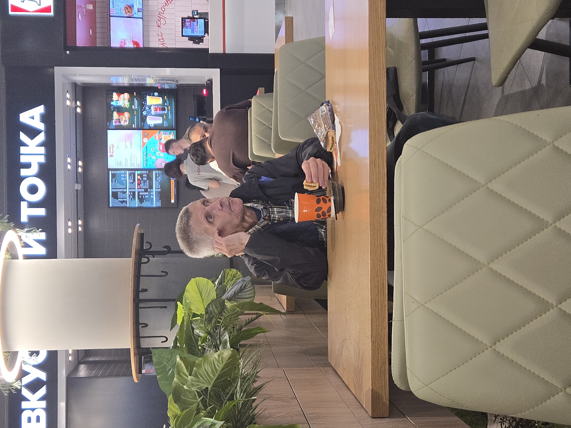 A man with gray hair is sitting at a table in a fast-food restaurant. He is wearing a black jacket over a plaid shirt and is holding a sandwich in his left hand, with his right hand touching his ear. An orange cup with black coffee beans is placed on the table in front of him, along with a wrapped snack. He appears to be looking off to the side. In the background, other people are visible, including a woman in a gray t-shirt with "MO" visible on it, and another person wearing a brown shirt. The restaurant has bright signage that reads "ВКУСНО И ТОЧКА" (Vkusno i Tochka), which is a Russian fast-food chain that replaced McDonald's. The environment includes wooden tables, light green chairs with a diamond-stitched pattern, and decorative plants. The overall scene suggests a casual dining experience in a modern fast-food establishment.