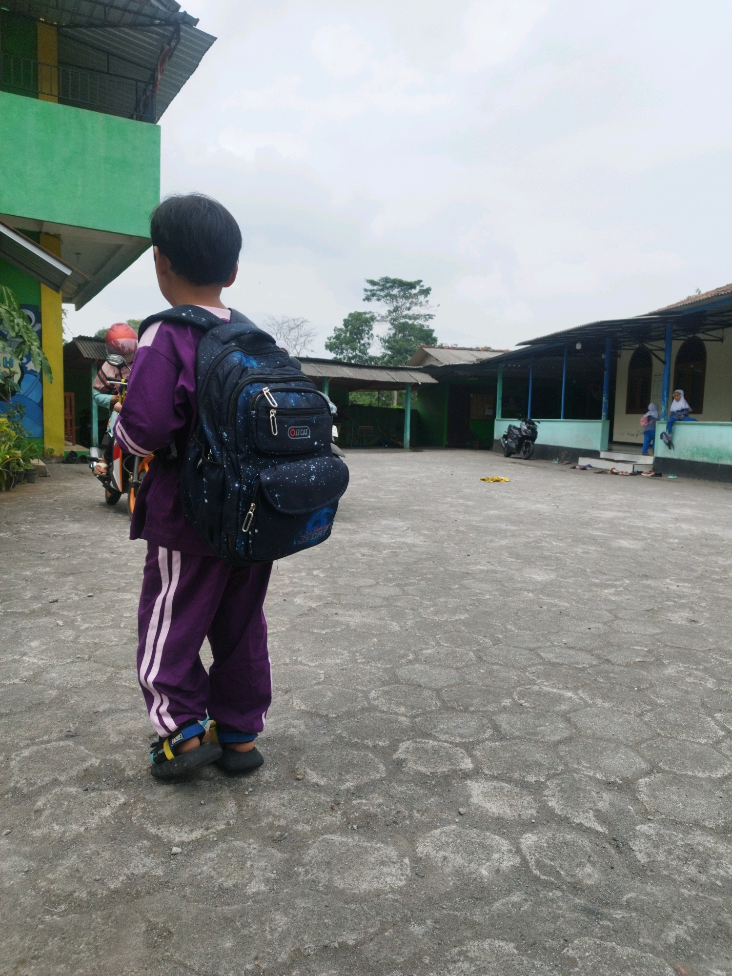 The image depicts a child wearing a purple uniform with white stripes on the pants and a matching shirt, carrying a dark blue backpack with a constellation pattern. The child is standing on a paved outdoor area, possibly a schoolyard, in what is identified as Hargobinangun, Indonesia. In the background, there is a green building with a second story. Further back, another building with a covered walkway and arched openings is visible. Two individuals wearing headscarves are seated on a ledge near this building. A motorcycle is parked in the vicinity. The ground is made of concrete pavers, and the sky is overcast. The scene appears to be a quiet, possibly educational, setting.