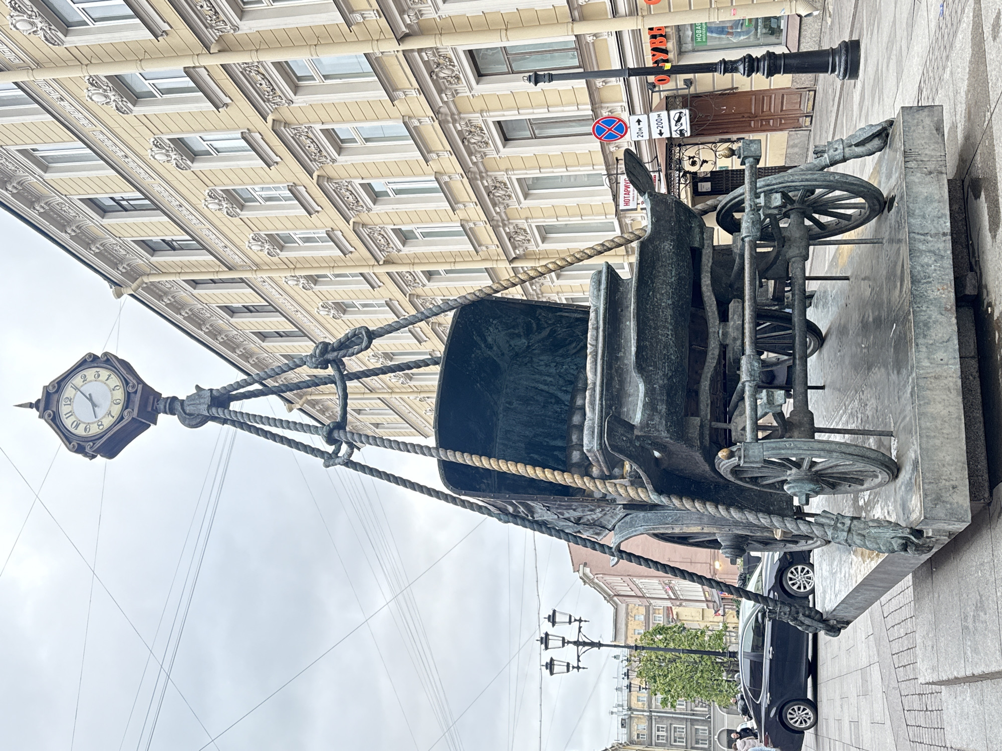The image depicts a bronze monument of a horse-drawn carriage with a clock attached to the front in Saint Petersburg, Russia. The sculpture stands on a paved sidewalk in front of a multi-story building with elaborate window frames. A street lamp is visible nearby, along with traffic signs. A black car is parked on the street. A person can be seen in the background wearing a face mask. The sky is overcast.