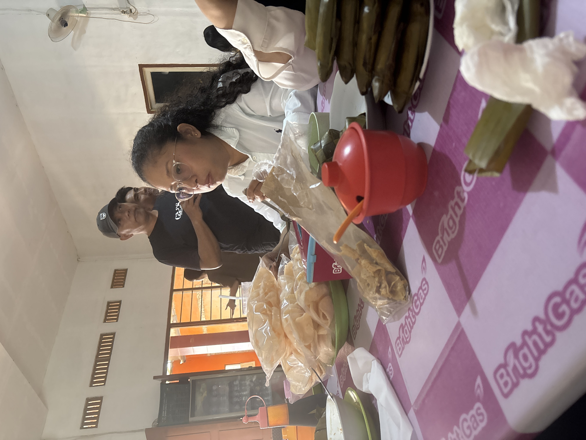 The image depicts a gathering at a table, likely in a community hall or a simple eatery in Merauke, Indonesia. The focus is on the food laid out on a pink and white checkered tablecloth. Visible food items include stacks of what appear to be rice cakes wrapped in banana leaves, bags of crackers, and what might be a bowl of crackers in a plastic bag. A small red condiment holder is also on the table.
Several individuals are present, though their interactions are not the primary focus. In the foreground, a woman wearing glasses and a white collared shirt is seated at the table, with her attention directed towards the food. Behind her, two men in casual attire, one wearing a baseball cap, are also present. The background reveals a room with white walls and ventilation grilles, suggesting a functional rather than decorative space. The overall impression is of a modest, communal meal setting in Merauke.