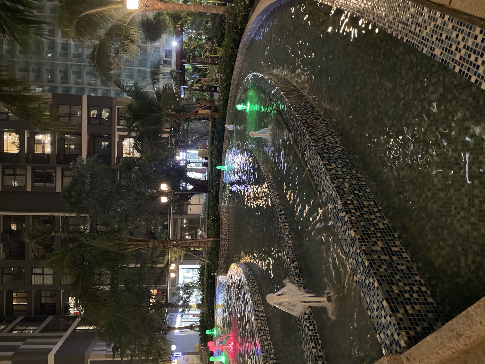 The image depicts a modern urban landscape at night in Hanoi, Vietnam, likely within a residential or commercial complex. In the foreground, a multi-tiered water fountain features several spouts. Some spouts produce white water jets, while others are illuminated by colored lights, casting green and pink hues onto the water. The fountain basin is lined with a mosaic of dark blue and white tiles.
Behind the fountain, palm trees and other greenery are visible. Further back, a series of tall apartment buildings dominate the skyline. These buildings are lit from within, with windows revealing interior lights, suggesting occupancy. The architecture is contemporary, characterized by large glass windows and dark facades.
In the middle ground, near the buildings and behind the greenery, there appears to be an outdoor recreation area, possibly a gym with exercise equipment. Several people are present, some standing and conversing, while others are near the playground equipment. Streetlights and decorative lighting are also present, illuminating pathways and accentuating the architectural features of the buildings. The overall atmosphere is that of a well-maintained, modern urban environment at night, with elements of leisure and residential living.