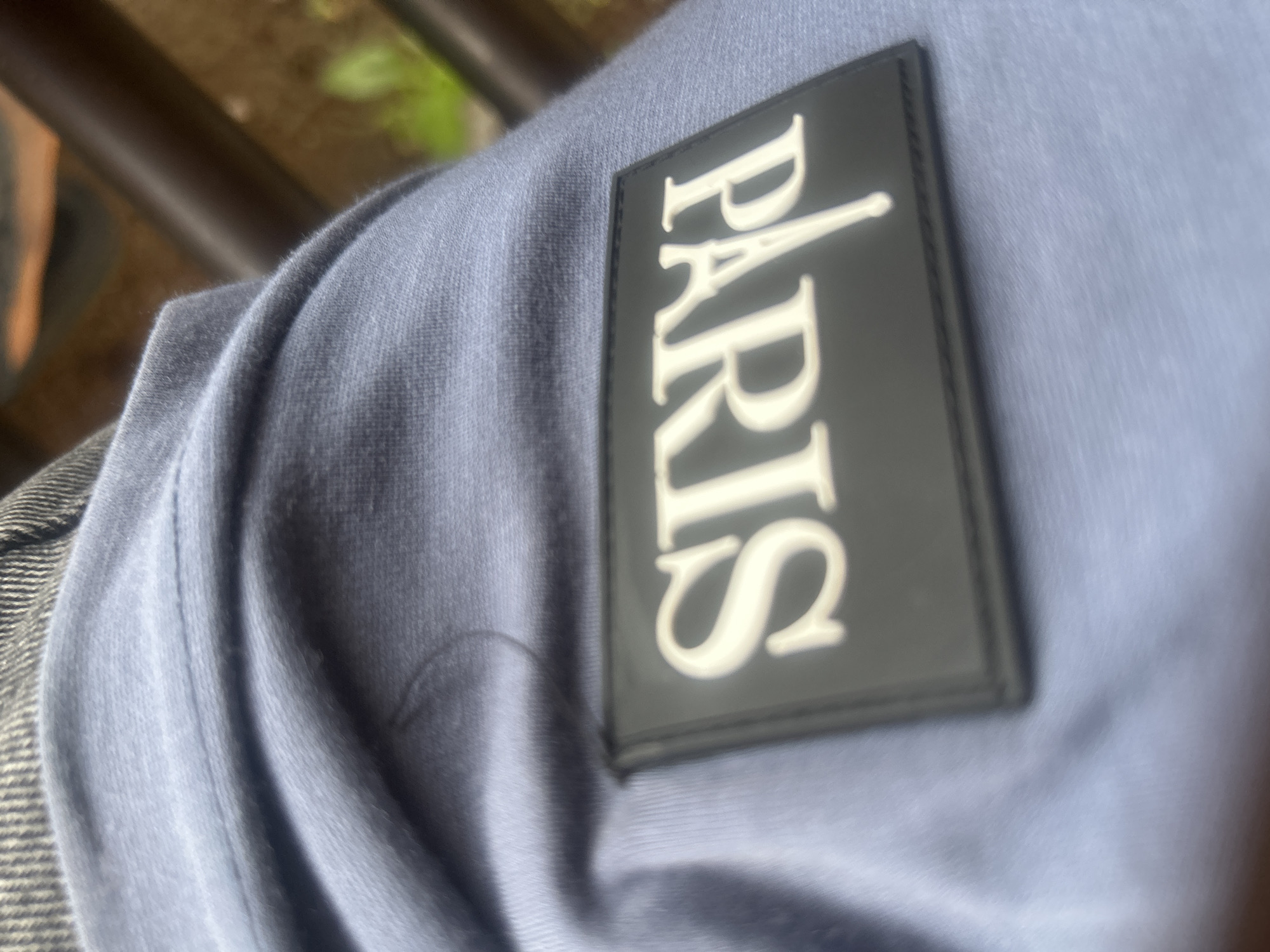 The image shows a close-up view of a piece of light blue fabric with a black patch stitched onto it. The patch has the word "PARIS" written in white, stylized capital letters. The fabric appears to be part of a garment, possibly a shirt or sweatshirt. The background is out of focus, but suggests an outdoor setting with brown bars and some green foliage. There is no discernible indication of the image being located in Ogbomosho, Nigeria.
