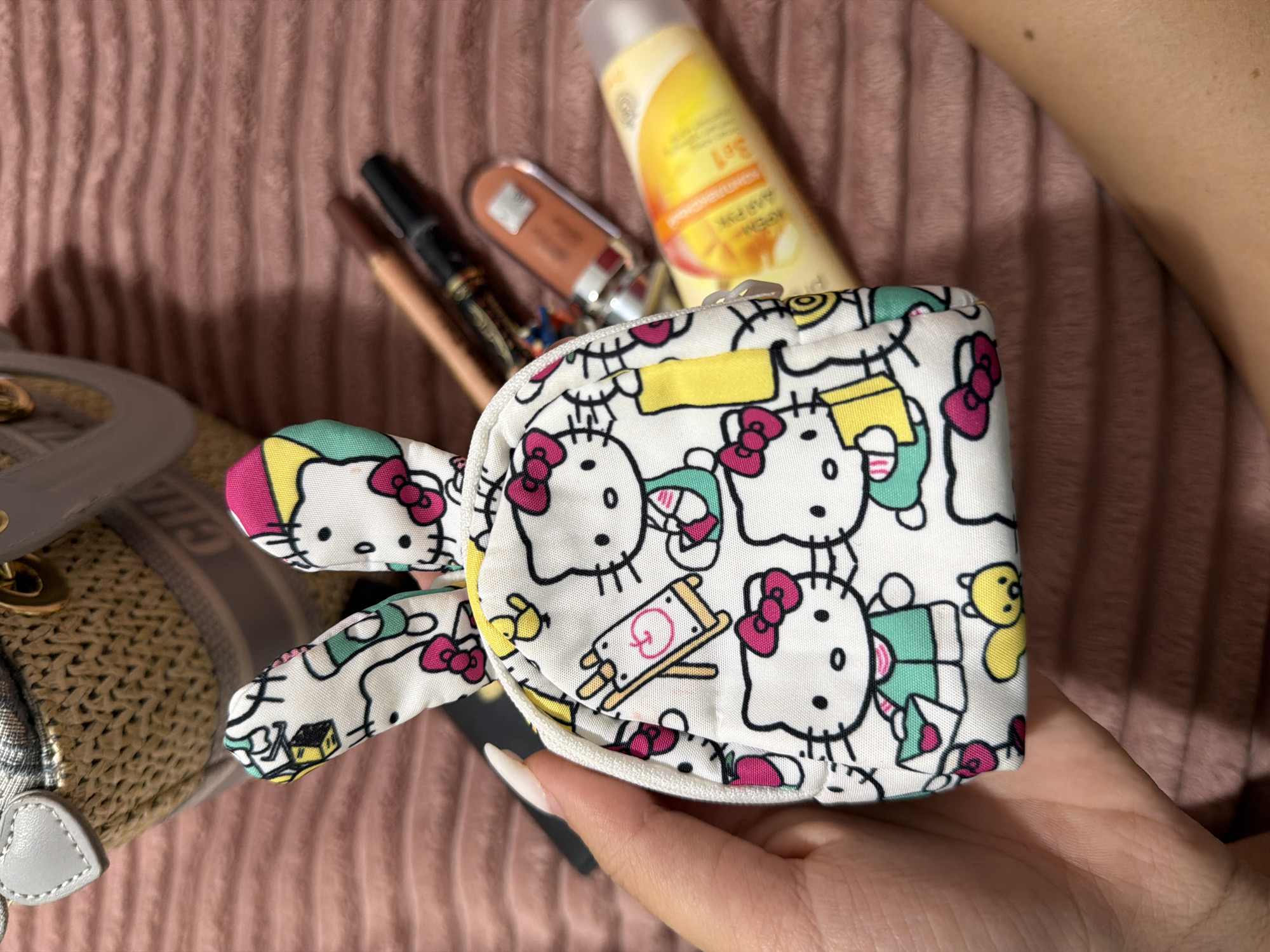 A person's hand, with a white-tipped, manicured fingernail, holds a small, white coin purse adorned with a Hello Kitty character print. The coin purse features Hello Kitty's face and ears, as well as other Hello Kitty-themed graphics. Behind the coin purse, several cosmetic items are visible, including two pencils, a tube of lip gloss, and a tube of sunblock. The items are resting on a pink, textured surface, possibly a blanket or fabric. In the upper left corner, a portion of a straw-like bag with gray leather trim is visible. There is no indication of the city of Digah, Azerbaijan in this image.