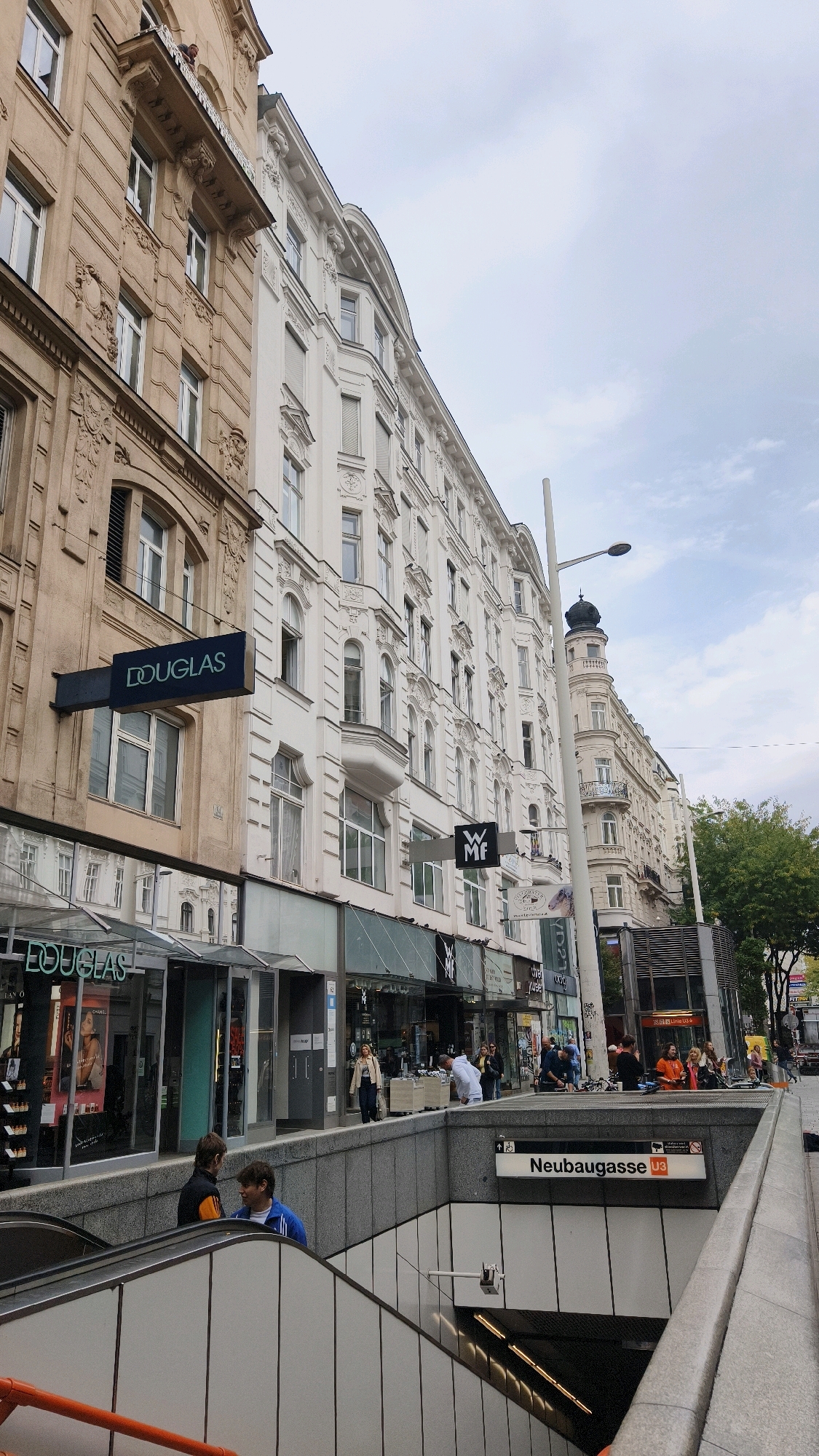 This is a street-level photograph of Neubaugasse U3 station in Vienna, Austria, on an overcast day. The perspective is from the top of the station entrance, looking down towards the escalators and up towards the street. On the left, a multi-story building with a beige facade is prominent, featuring ornate architectural details and balconies. A dark sign for "DOUGLAS" is affixed to its facade. Adjacent to it is a white building with similar classical architectural elements and bay windows. Further down the street, more buildings line the sidewalk, some with commercial storefronts. Several people are visible on the street and sidewalk, including a woman in a beige jacket, a man in a white shirt, and groups of people conversing. The street is relatively busy with pedestrian traffic. The entrance to the Neubaugasse U3 subway station is the focal point in the foreground, with gray stone walls and white tiered panels leading down to escalators. A station sign, "Neubaugasse U3," is clearly visible above the entrance. In the background, a street lamp and trees can be seen.