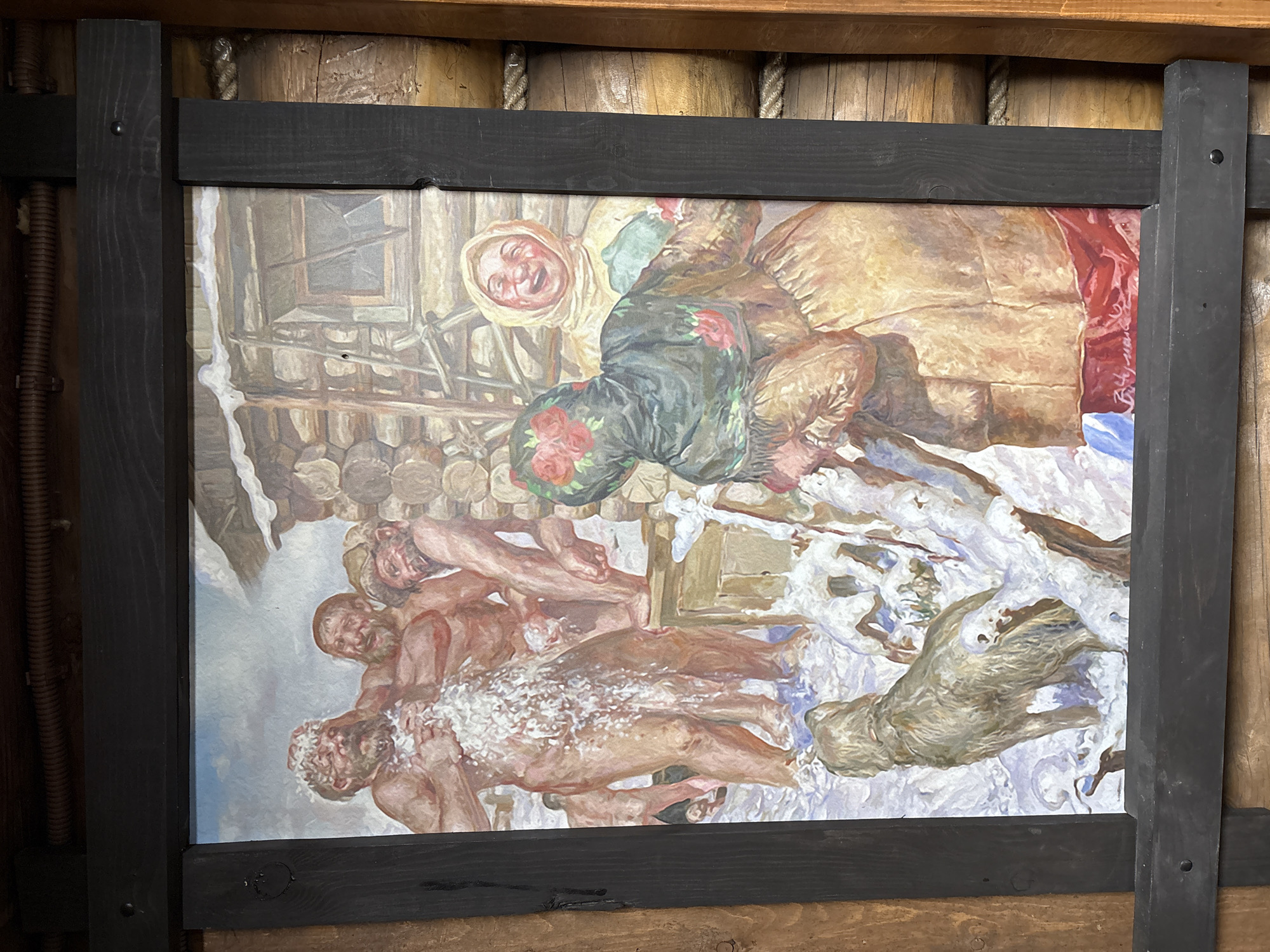 The image shows a painting displayed in a dark wooden frame. The painting depicts a scene with a group of people in a snowy environment. In the upper portion of the painting, a woman with a joyful expression is visible, wearing a white headscarf and possibly a tan-colored coat. She appears to be looking down at a group of naked men lying in the snow. The men are pale and some are covered in snow. One animal is also in the bottom right corner of the painting. The painting is on display in the City of Prikamskii, Russia.