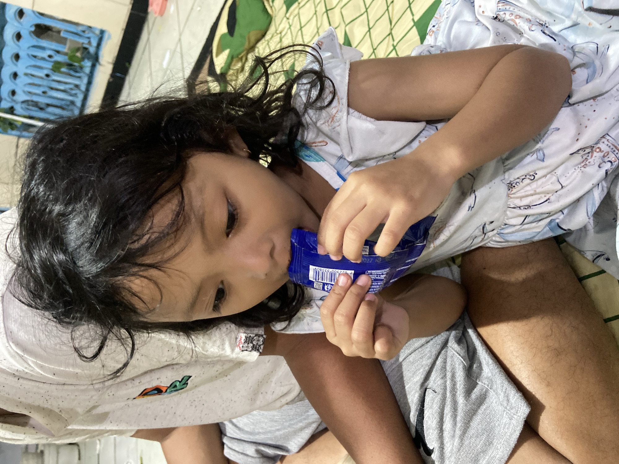 The image shows a young girl in Gelam, Indonesia, lying down on a patterned mattress and possibly a bed. She has long, dark, curly hair and is wearing a light-colored dress with a pattern of blue and white illustrations. She is holding a blue package, possibly of snacks or cookies, and appears to be eating from it. Her facial expression is relaxed, suggesting a moment of contentment.
The setting appears to be indoors, possibly a bedroom, given the presence of a mattress and what looks like a wall or a partition in the background. There are hints of a blue plastic rack or shelf in the upper left corner, and a camouflage-patterned object visible on the mattress to the left of the girl. The lighting suggests it could be daytime, though the exact time of day is not definitively ascertainable.
On the blue packaging, there is visible text, including a barcode and some alphanumeric characters. Some of the visible text includes "0053309", "0 23500", and what appears to be "2027 N". The girl's legs are also visible, with one leg bent and resting on top of the other. She is wearing grey shorts or sweatpants. The overall mood is casual and intimate, capturing a simple moment of childhood enjoyment.
