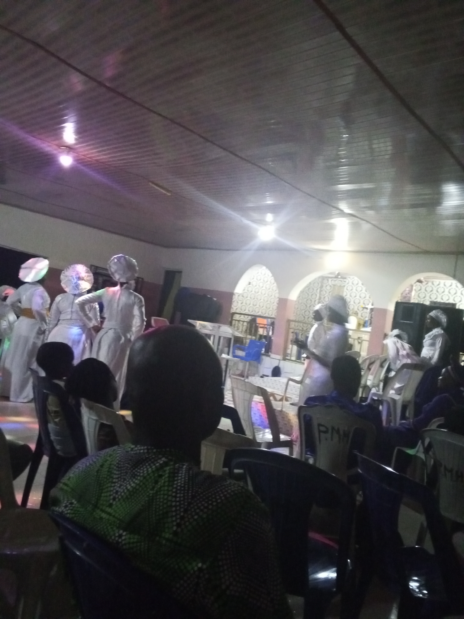 The image shows an indoor event, possibly a religious gathering, in Lokoja, Nigeria. Several people wearing white robes and elaborate headwear are standing on a slightly raised platform. They appear to be performing or presenting something. In front of them, several people are seated in chairs, watching the event. The interior has a decorated wall with arches and a ceiling with lights. Some chairs have markings on them, possibly labels or names. The lighting is somewhat dim, creating a subdued atmosphere.
