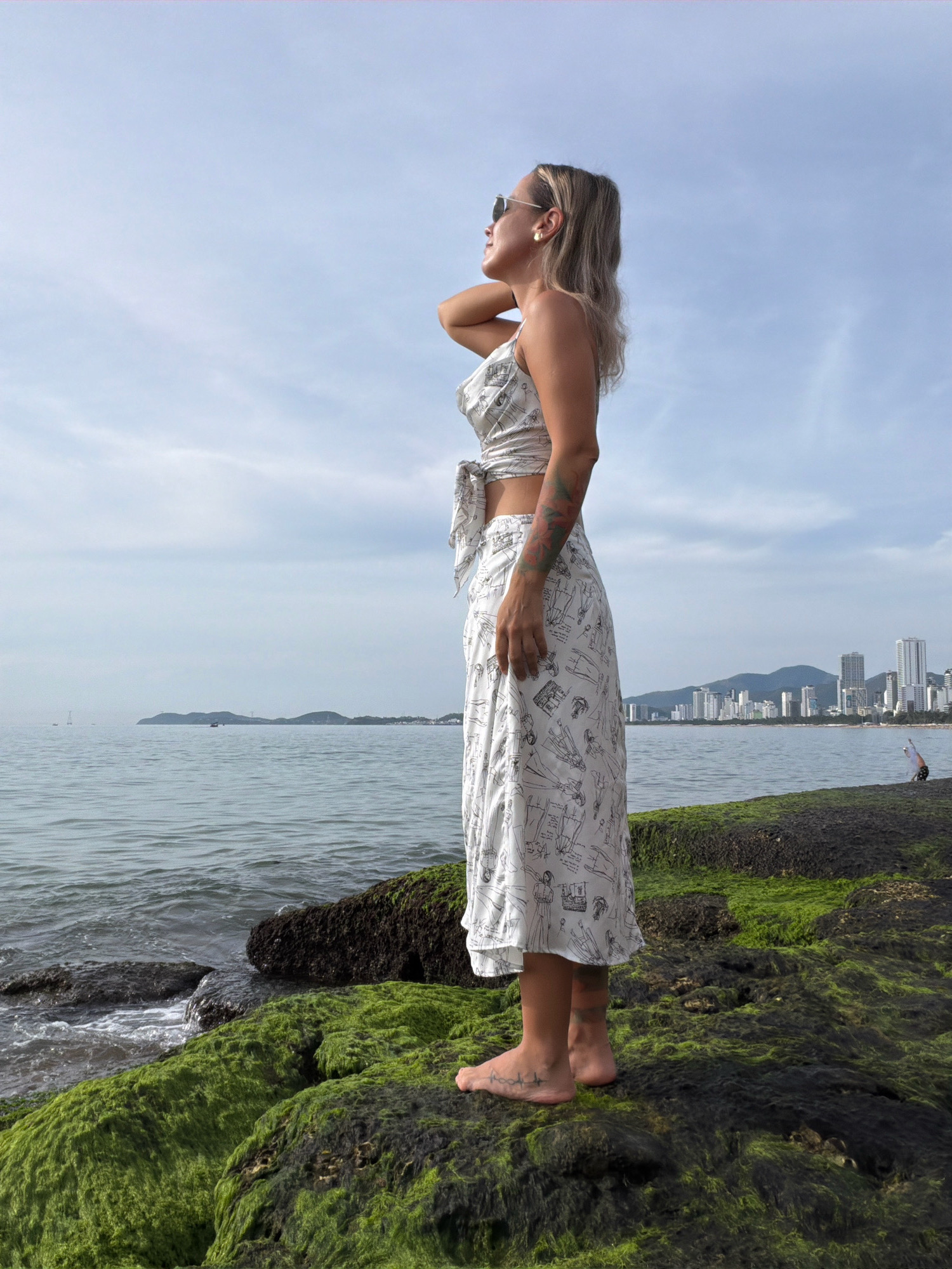 A woman stands on moss-covered rocks by the sea in Nha Trang, Vietnam, looking out at the water. She has blonde hair pulled back and is wearing sunglasses, a patterned two-piece outfit, and a long skirt. The ocean is calm with a few boats in the distance. A city skyline with tall buildings is visible across the water. The sky is partly cloudy.