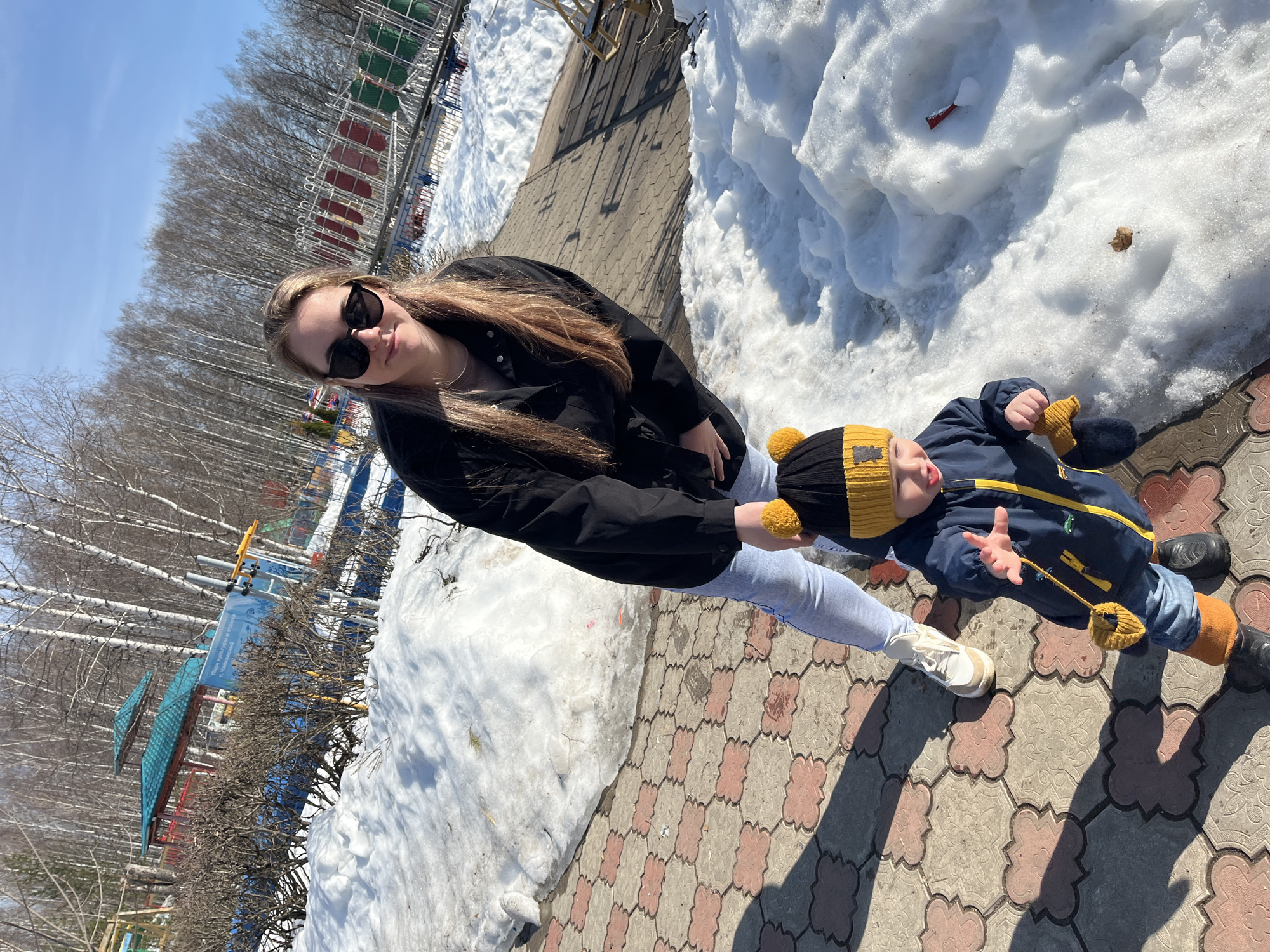 The photo depicts a young woman and a child on a paved walkway in Chaikovsky, Russia, likely in a park or recreational area. The woman is wearing sunglasses, a black jacket, and light-colored jeans and sneakers. The child is dressed in a blue winter coat, yellow and black hat, and boots. Patches of snow are visible alongside the walkway, suggesting it's late winter or early spring. Trees line the background. The woman appears to be holding the child's hand.