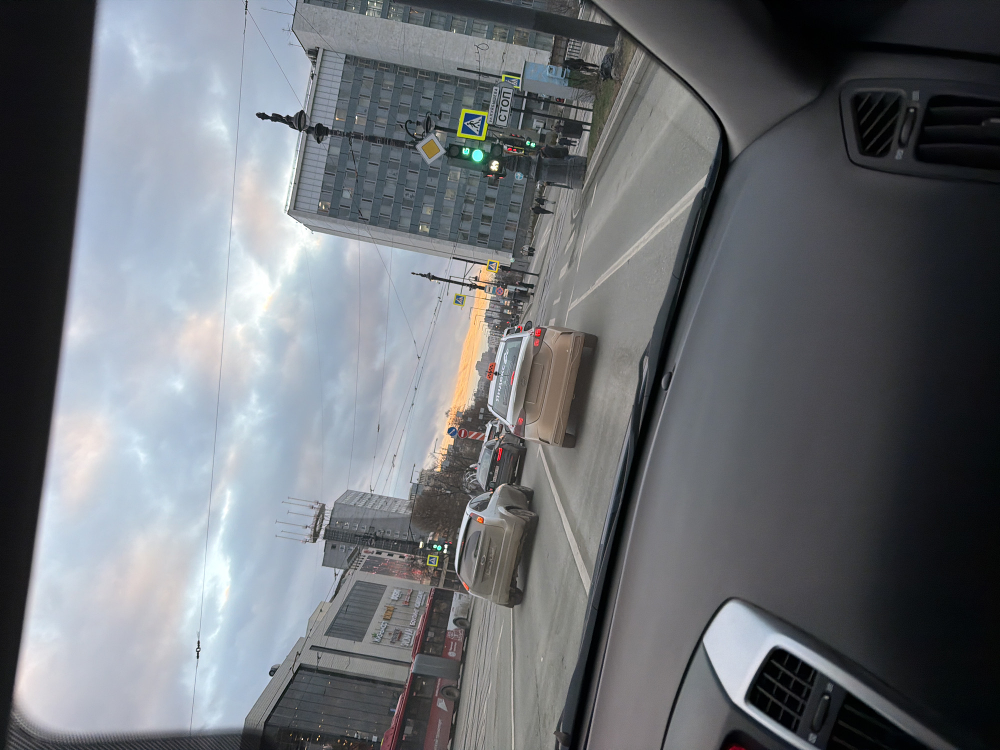 The view is from inside a car, looking out onto a street in Perm, Russia. Traffic is moving along the road. A tall, Soviet-era building is visible on the left, with smaller commercial buildings at its base. A red bus is in the traffic. Street lights and traffic signals are present. The sky is overcast.