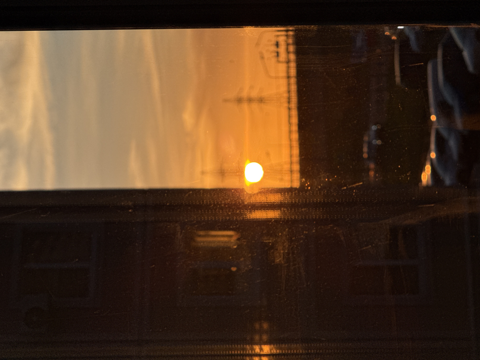 The image, taken through a dirty window, depicts a sunset reflecting off of buildings in Chaltyr', Russia. The sun is low on the horizon, with a yellow glow. Power lines are visible in the reflection. Buildings are visible both through the window and in the reflection. Cars can also be seen in the reflection, partially obscured by streaks on the glass.