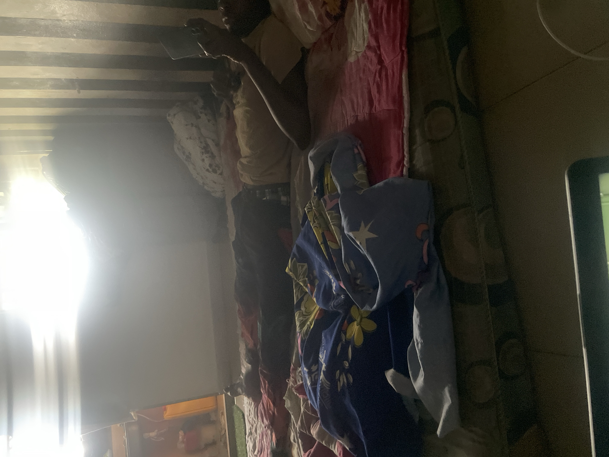 The image depicts an indoor scene, likely a bedroom, with a person lying on a bed covered with colorful blankets. The room appears dimly lit, with a bright light source emanating from the upper left, possibly a window or an artificial light, creating a strong glare. The person is lying on their back, with one arm raised, holding a mobile phone. The bed is cluttered with blankets and pillows. In the background, there are vertical stripes on the wall, suggesting a textured wallpaper or corrugated material. The floor appears to be tiled. The overall atmosphere suggests a casual or domestic setting. The location is identified as Ihiagwa, Nigeria.