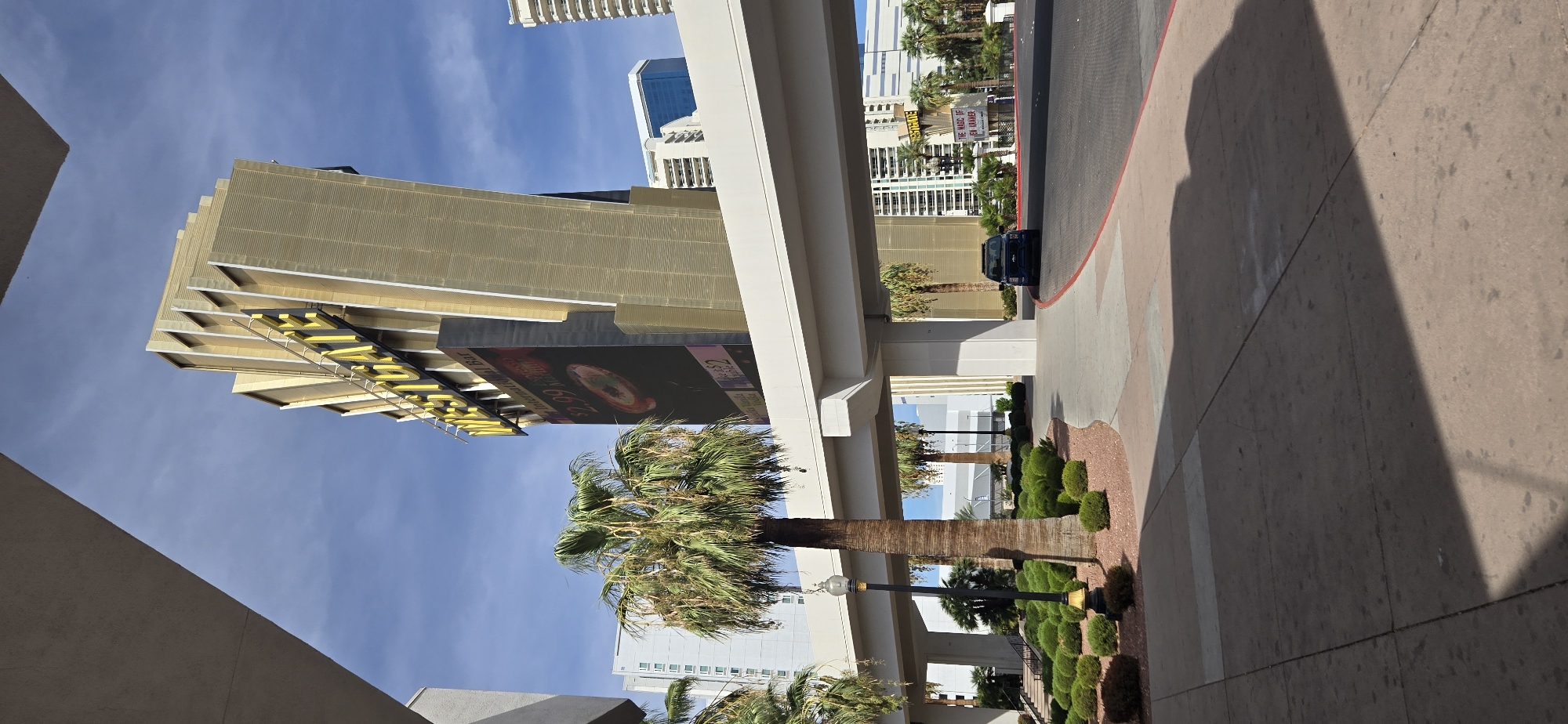 The image depicts the exterior of the Westgate Las Vegas, a resort and casino located on the Las Vegas Strip in Paradise, Nevada, USA. In the foreground, a paved walkway and a landscaped area with palm trees and manicured bushes are visible. A dark-colored SUV is driving on the street under an elevated walkway. In the background, the prominent Westgate Las Vegas sign, illuminated in yellow, stands tall against a blue sky with scattered clouds. A large digital billboard displays advertisements, including a promotion for "$2.99 Martinis." Other buildings, including high-rise hotels, are visible in the distance. The scene appears to be an outdoor public space, likely a street or sidewalk area adjacent to the resort.