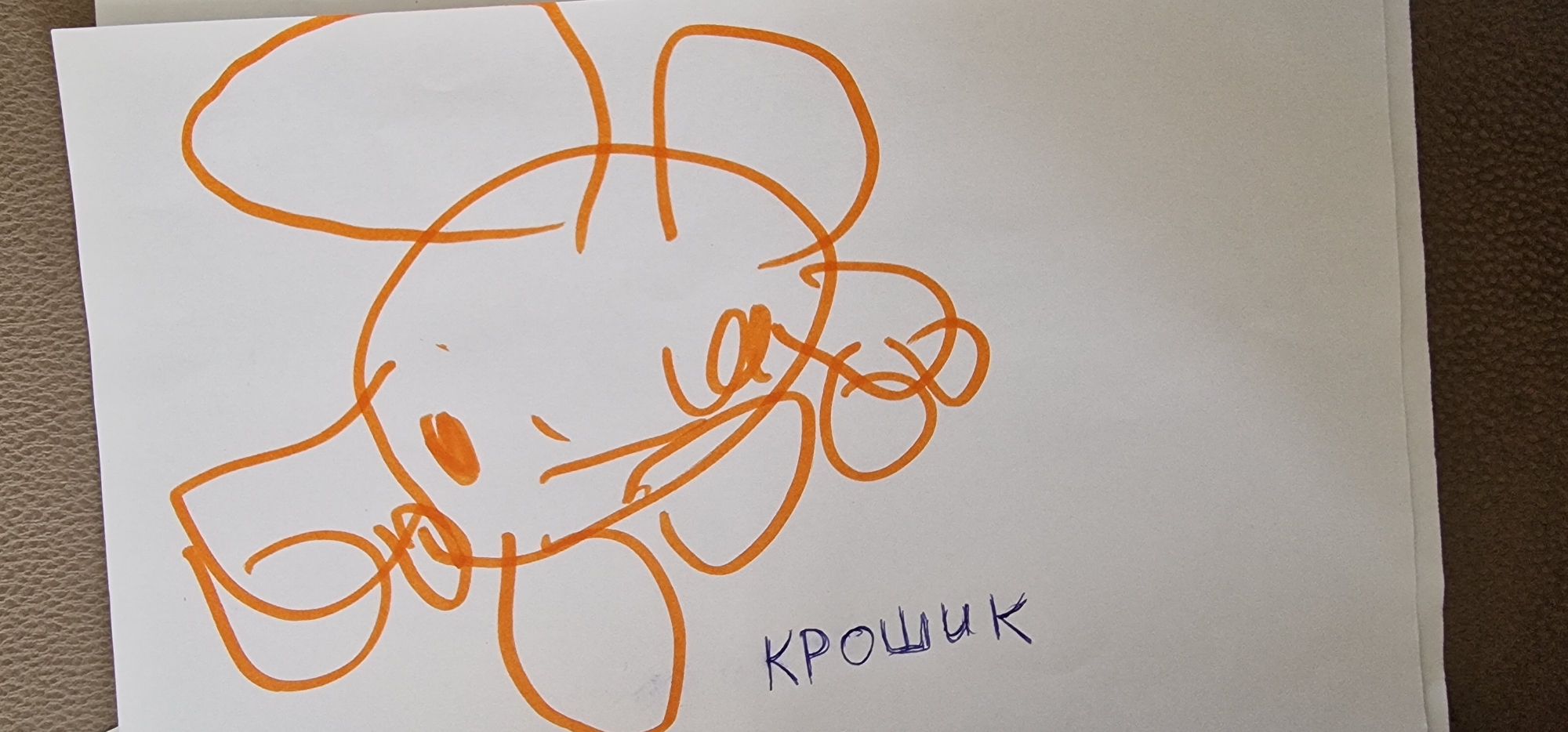 The image shows a child's drawing of an elephant with large, curly ears and a trunk. The drawing is in orange marker on a white piece of paper. Below the drawing, the word "КРОШИК" is written in blue ink. The paper is placed on a textured surface that appears to be a brown couch or carpet. There is no identifiable location in the city of Daugavpils, Latvia.