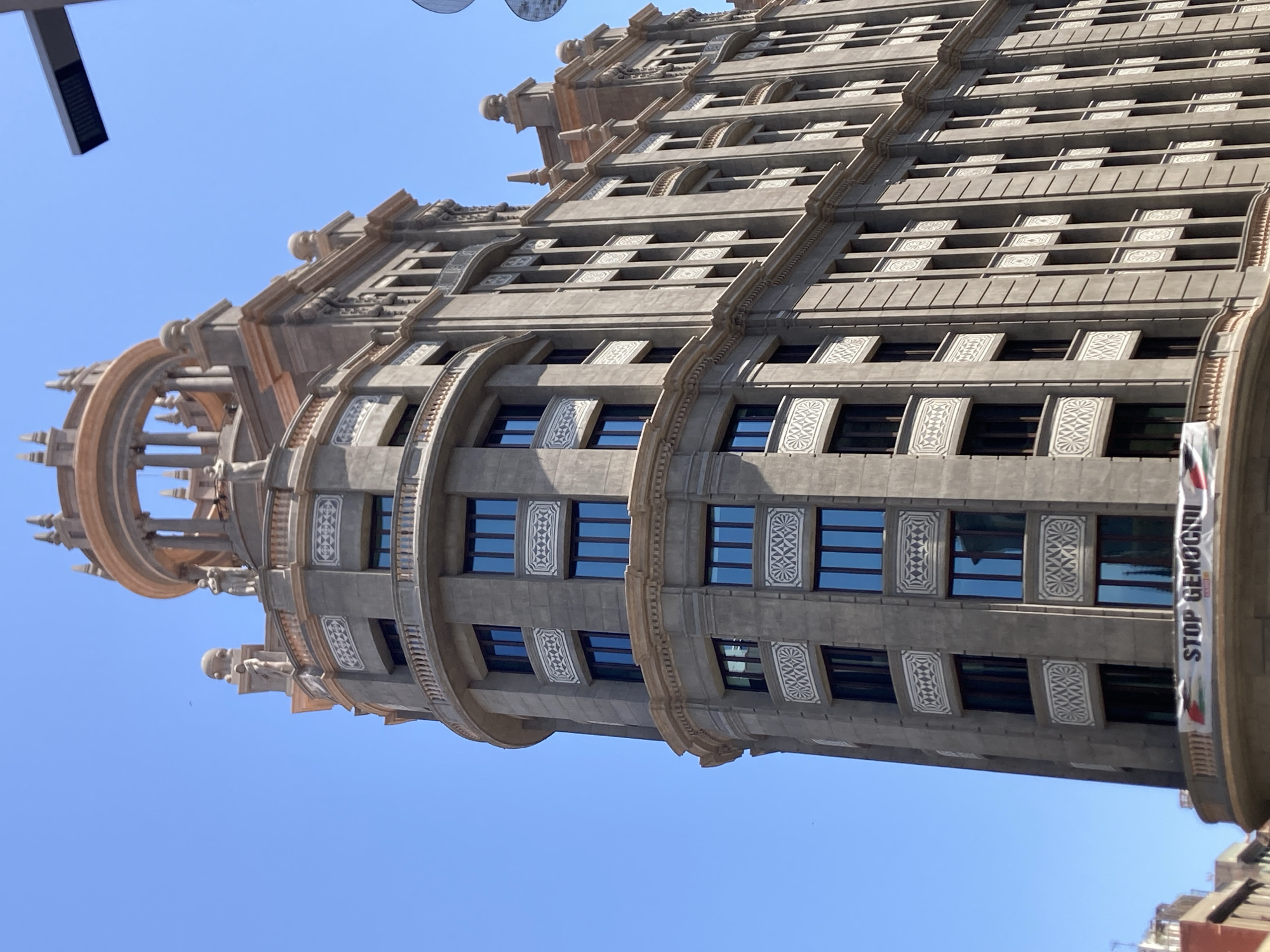 The image shows a historic building in Barcelona, Spain, with a banner that reads "STOP GENOCIDI". The building is tall and ornate, with multiple stories and balconies. The banner is hanging from a balcony on the lower part of the building, and it is made of fabric with a checkered pattern of red, white, black, and green stripes on the sides, and white in the middle with black lettering and a logo. The sky is clear and blue, and the sun is shining. The building's architecture features curved facades, decorative elements, and numerous windows. The banner is partially visible, extending across the width of the balcony. The building appears to be made of concrete or stone, with a greyish hue. The overall scene depicts a juxtaposition of architectural grandeur and a socio-political message.