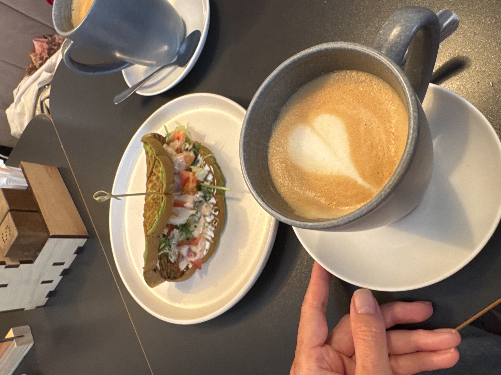 A high-angle shot depicts a table scene, with a focus on culinary and beverage items. On the dark-colored table are two light-colored saucers, each holding a gray-blue cup filled with coffee or latte, indicated by the milk foam art. The saucer to the right is slightly elevated by a partially visible hand. A white plate holds a half-eaten pita or taco-like food item, secured with a toothpick. To the left, a decorative wooden box sits next to the plate. The background shows a hint of an interior, possibly a cafe or restaurant. Due to the lack of recognizable landmarks, precise location is not possible.