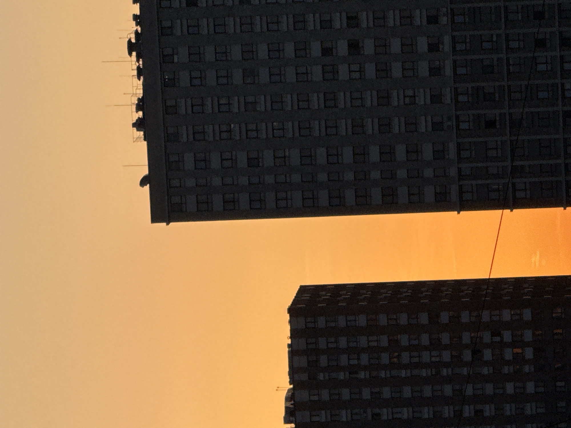 The image depicts two high-rise buildings against a bright orange sky. The buildings are dark in color and appear to have many windows. The sky is uniform in color. The location is most likely urban.