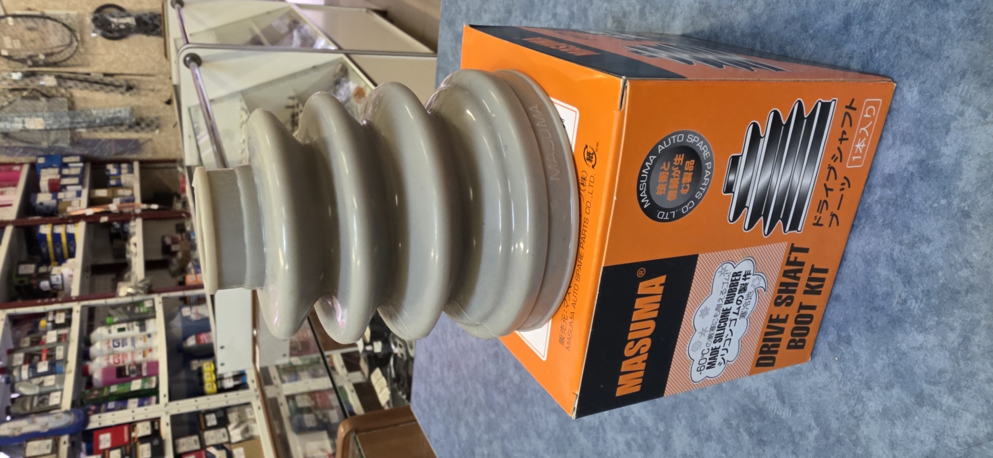 The image shows an orange cardboard box with a gray rubber boot resting on top. The box is labeled "MASUMA" in large orange letters and also contains text in Japanese and English. The English text reads "DRIVE SHAFT BOOT KIT" and "MADE SILICONE RUBBER". The rubber boot appears to be a new car part, possibly a CV joint boot. The box is sitting on a blue surface, which is likely a table. In the background, shelves filled with various items are visible, suggesting a retail store or warehouse environment. The overall impression is of a product display. The image does not contain any information that allows for the determination of its location in Kryvyi Rih, Ukraine.
