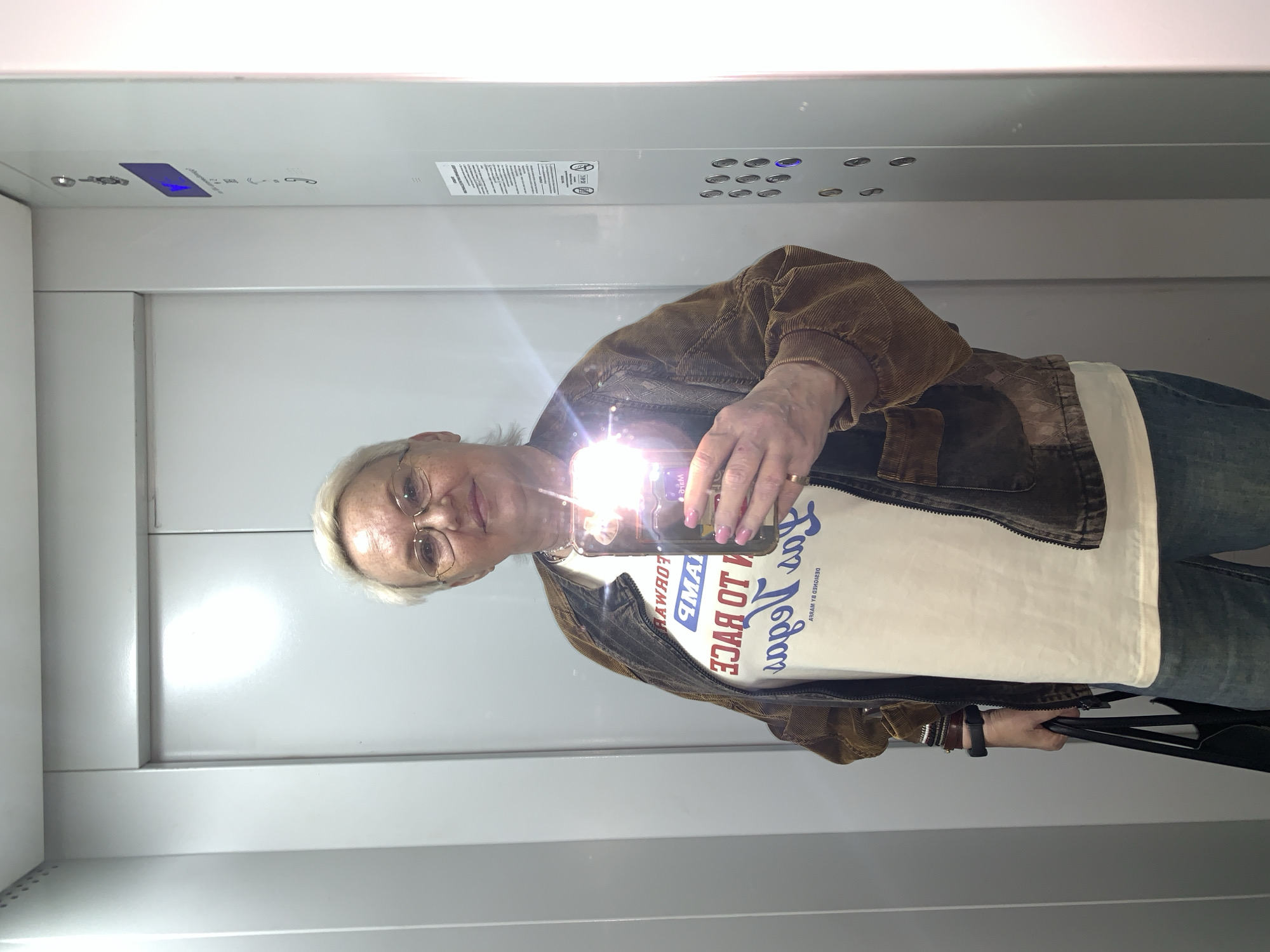 An older woman with short, light-colored hair and glasses is captured in a mirror selfie inside an elevator in Chaikovsky, Russia. The image is rotated 90 degrees counter-clockwise. She holds a smartphone, likely with its flash on, creating a bright glare that obscures part of the phone's screen and her chest. She wears a brown corduroy jacket over a white t-shirt, blue jeans, and carries a black bag in her left hand. A watch or fitness tracker is visible on her left wrist, and she wears a ring on her right hand. Her expression is neutral with a slight smile.
The elevator interior features sleek, light gray walls. A control panel on the upper right has several round, illuminated buttons, with some showing a blue glow, indicating activated functions or selected floors. Below these, there's a smaller set of buttons, likely for emergency and door controls, and various standard pictograms including a bell, a telephone, and accessibility symbols. A white label with fine print is also affixed to the panel.
The text on the woman's white t-shirt is visible in reverse due to the mirror reflection. It appears to read "DESIGNED BY HYP" and phrases like "FOR GOOD", "TO RACE", and "FORWARD". The scene suggests a casual moment of self-documentation within a modern, well-maintained indoor environment.