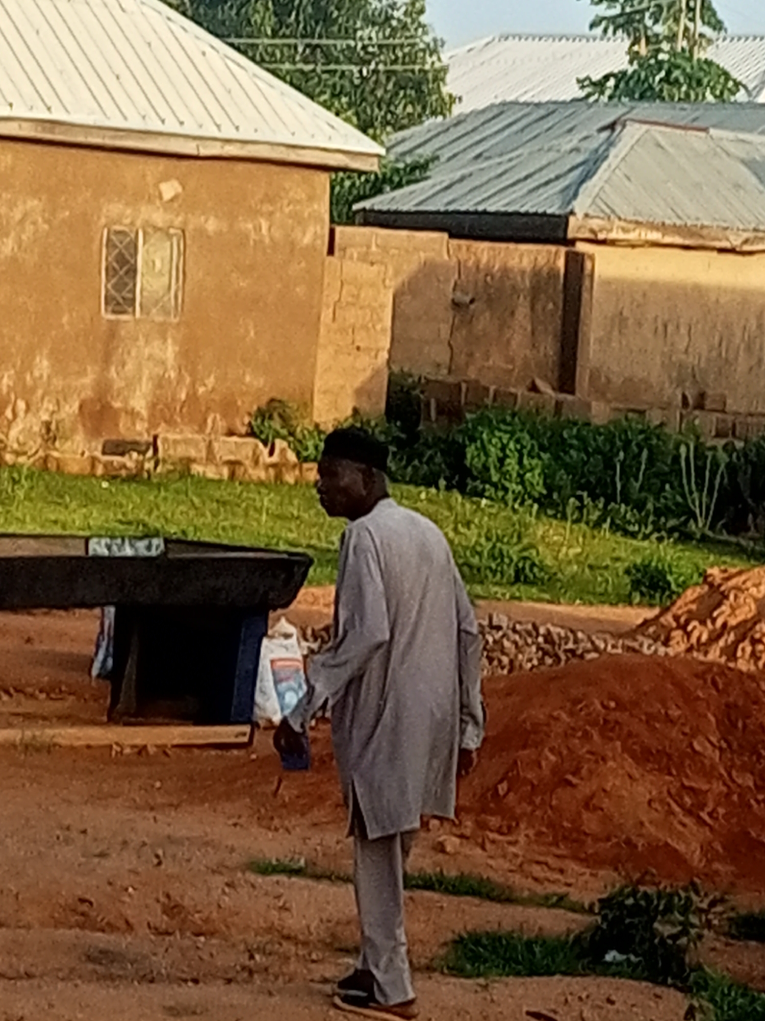 A man stands outdoors, facing away from the camera and looking to the right. He is wearing a gray tunic and pants, and a black skullcap. He is standing on a dirt ground, in front of a pile of red dirt or soil. In the background, there are two buildings. The building on the left is made of mud and has a white roof. It has a single window. The building on the right is made of bricks and also has a gray metal roof. There is green grass and some plants growing in front of the buildings.