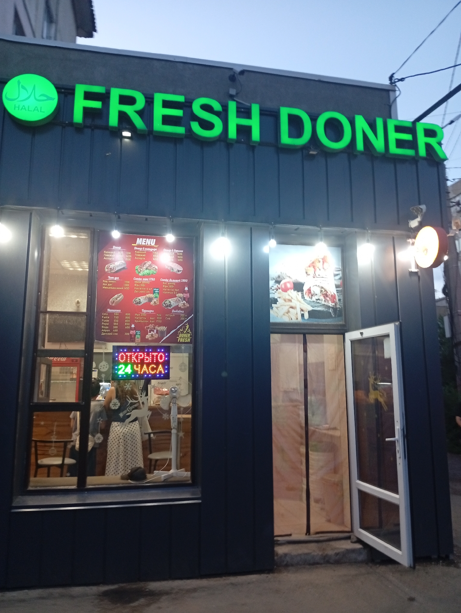The image shows the exterior of a "FRESH DONER" establishment in the city of Oral, Kazakhstan. The building is dark gray with large windows. Above the windows, a bright green neon sign reads "FRESH DONER" with a green circular logo on the left that says "HALAL" in Arabic script. Inside the window on the left, a menu is displayed. A neon sign in the window on the right flashes "ОТКРЫТО 24 ЧАСА" which translates to "OPEN 24 HOURS." Two people are visible inside the establishment. One person, appearing to be a woman with a patterned skirt, is standing behind a counter, and another person is partially visible in the background. The ground in front of the establishment appears to be concrete. A partially open glass door is visible on the right side of the building. The sky is overcast.