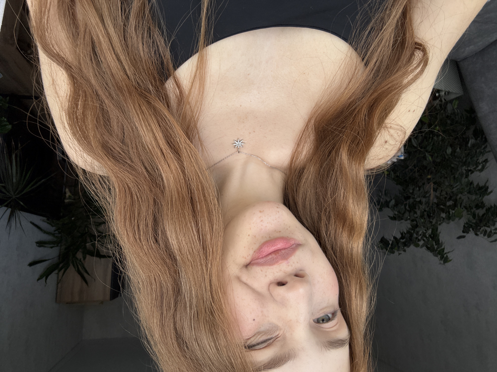 This close-up, vertically oriented image captures a young woman looking directly at the viewer with a playful expression. Her head is tilted, and she winks with her left eye while her right eye, which is light-colored (blue-green), remains open. Her lips are slightly parted, revealing a soft pink color, and her fair skin is adorned with numerous freckles across her nose and cheeks. Long, wavy, reddish-brown hair frames her face and shoulders, cascading around her.
She is wearing a simple black top, and a delicate silver chain necklace with a small, star-shaped pendant featuring a tiny gemstone rests on her upper chest/collarbone area. The background is softly blurred, showing dark green foliage from plants, suggesting an indoor or semi-outdoor setting like a balcony or patio. The lighting appears natural and gentle, creating a soft glow on her skin. The overall mood is casual and friendly. There is no visible text in the image. The location is Naberezhnye Chelny, Russia, though no specific visual cues to this city are present within the frame beyond what could be a common domestic setting.