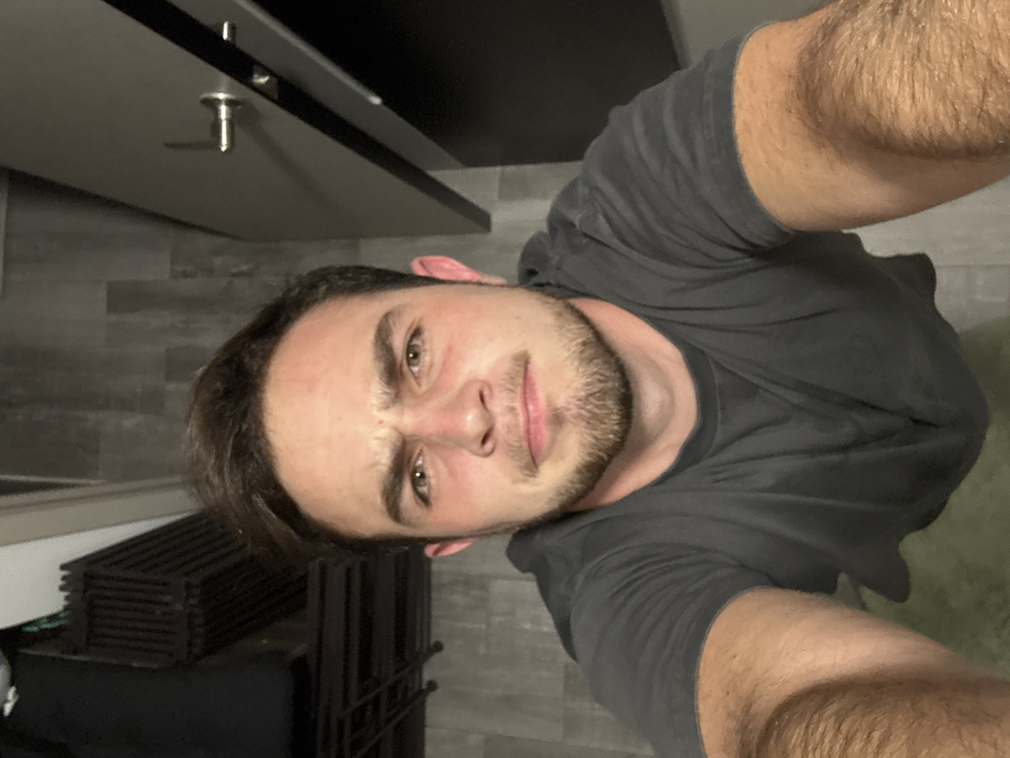 A man with short brown hair and a beard is posing for a selfie indoors. He's wearing a dark grey t-shirt. Behind him, there is a kitchen cabinet and a grey tiled wall. A black object, possibly a rack, is visible on the left side of the frame. The image does not allow for a determination of location in San Bernardino Tlaxcalancingo, Mexico.