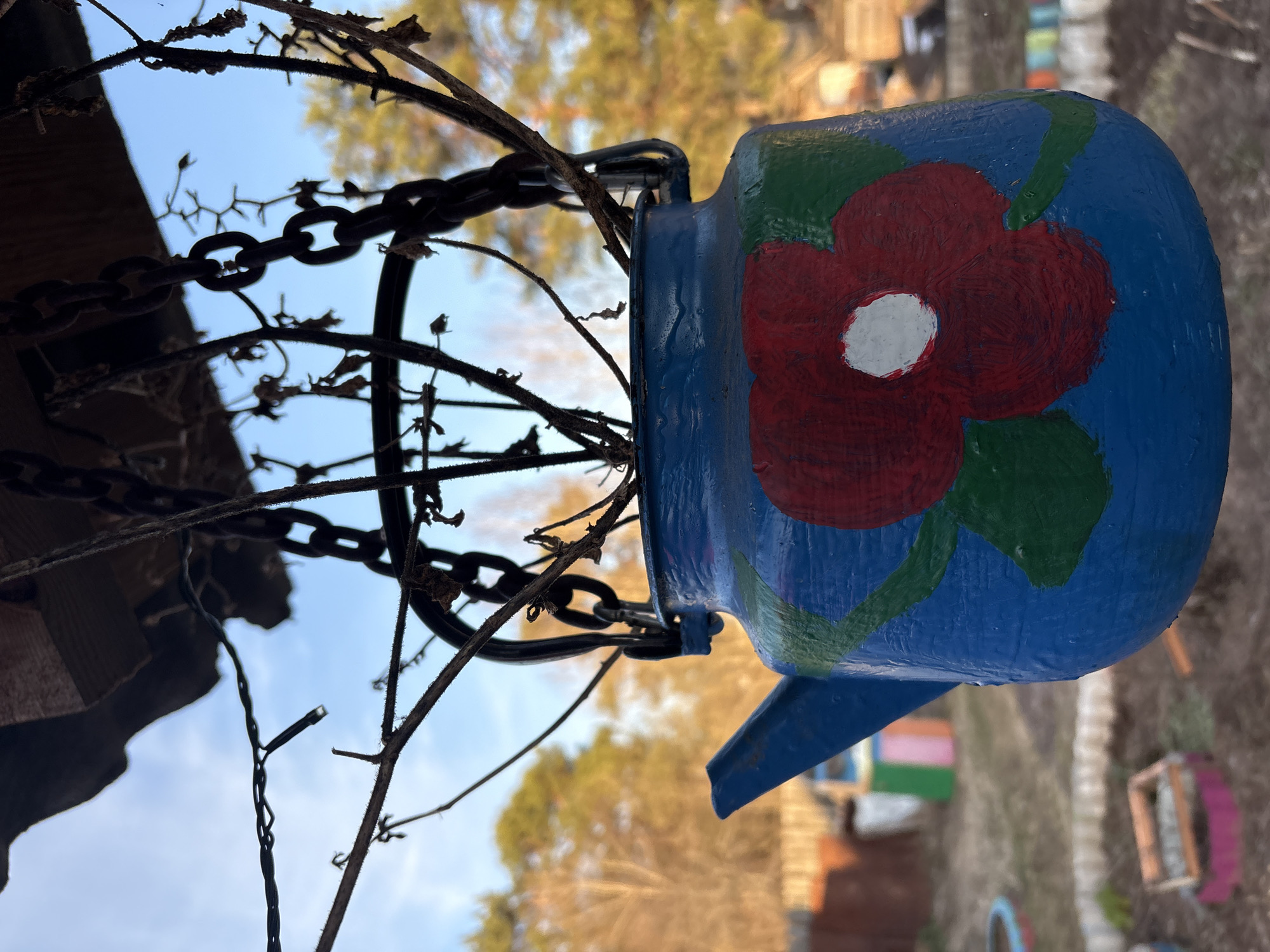 This close-up, rotated image captures a decorative outdoor scene, likely in a garden or yard in Chaikovsky, Russia, during daytime. The primary subject is a vibrant blue teapot, repurposed as a hanging ornament or planter. It is hand-painted with a prominent red flower featuring a white center, surrounded by green leaves.
The teapot is suspended by a dark, metallic chain, which itself is intertwined with a network of dry, bare branches or vines. These elements hang down from a dark, unseen structure, part of which, resembling a wooden beam or roof edge, is visible in the upper left. A delicate string of small lights is also visible, woven among the branches.
The background reveals a bright, clear blue sky with minimal cloud cover in the upper half. Below the sky, out-of-focus golden and brown foliage of trees suggests an autumn setting. The lower background shows hints of a rustic outdoor space, including a fence and various colorful objects, possibly painted tires or pots, scattered on the ground, contributing to a cheerful, handcrafted aesthetic. The lighting is bright and natural, indicating a sunny day. No visible text is present in the image.
